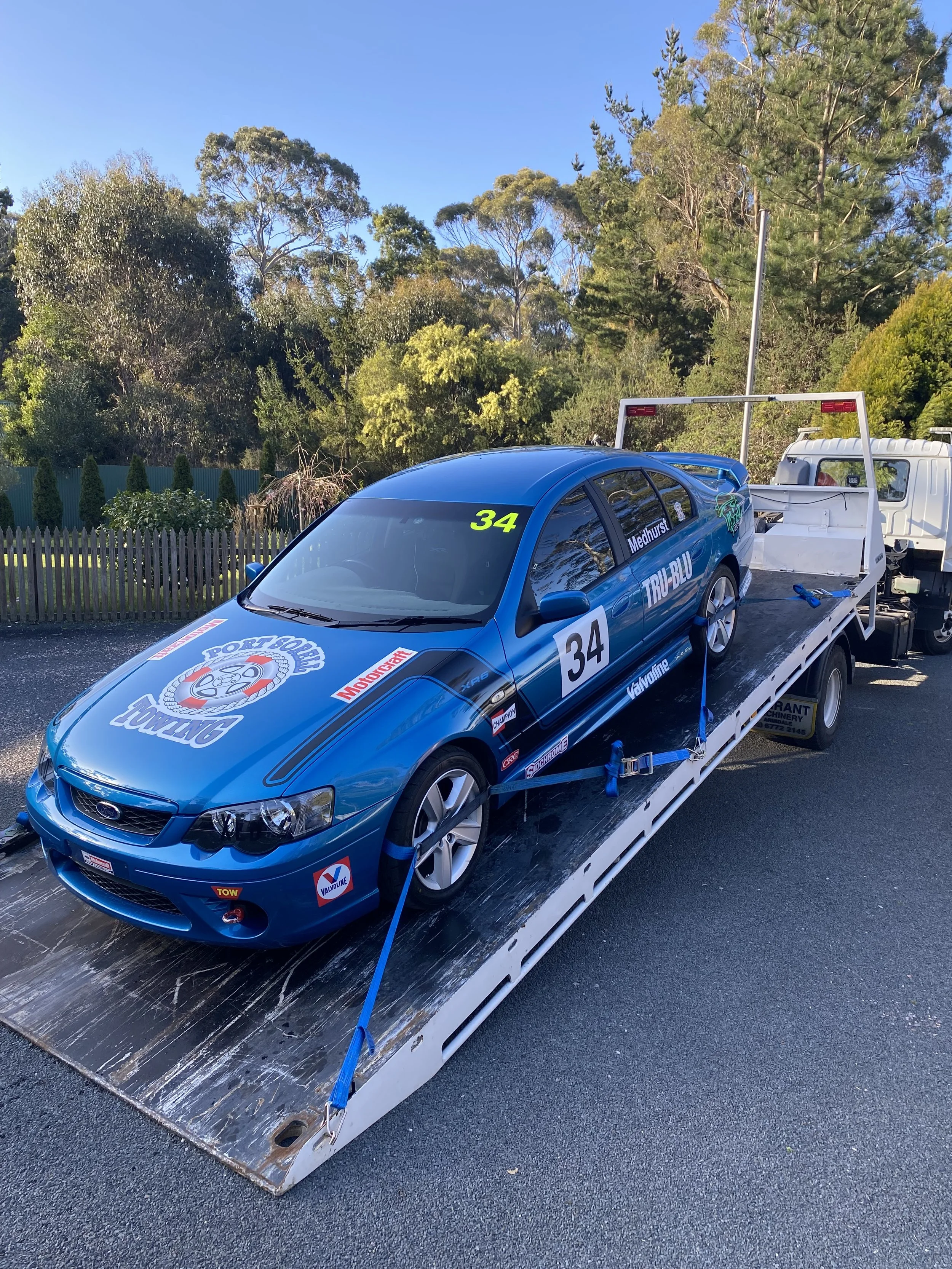 A blue race car on a flatbed truck, with racing decals and the number 34, parked outdoors with trees and sky in the background.