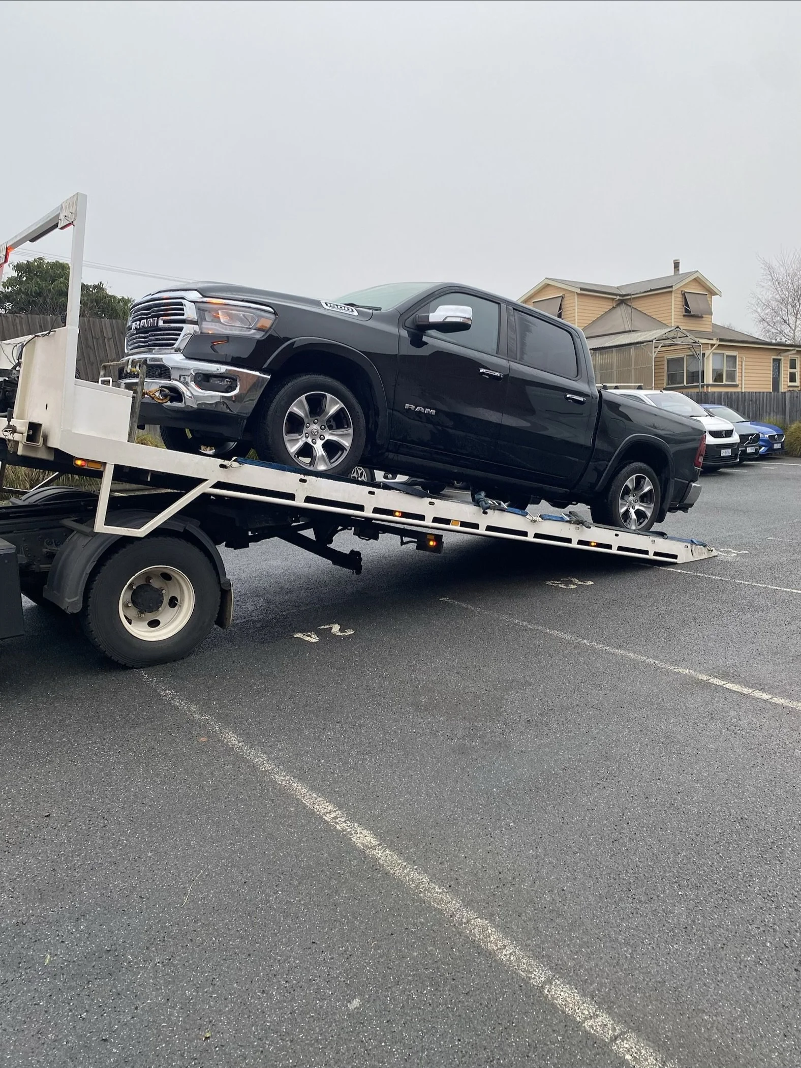 Black pickup truck on a flatbed tow truck in a parking lot, with other parked cars and residential houses in the background.