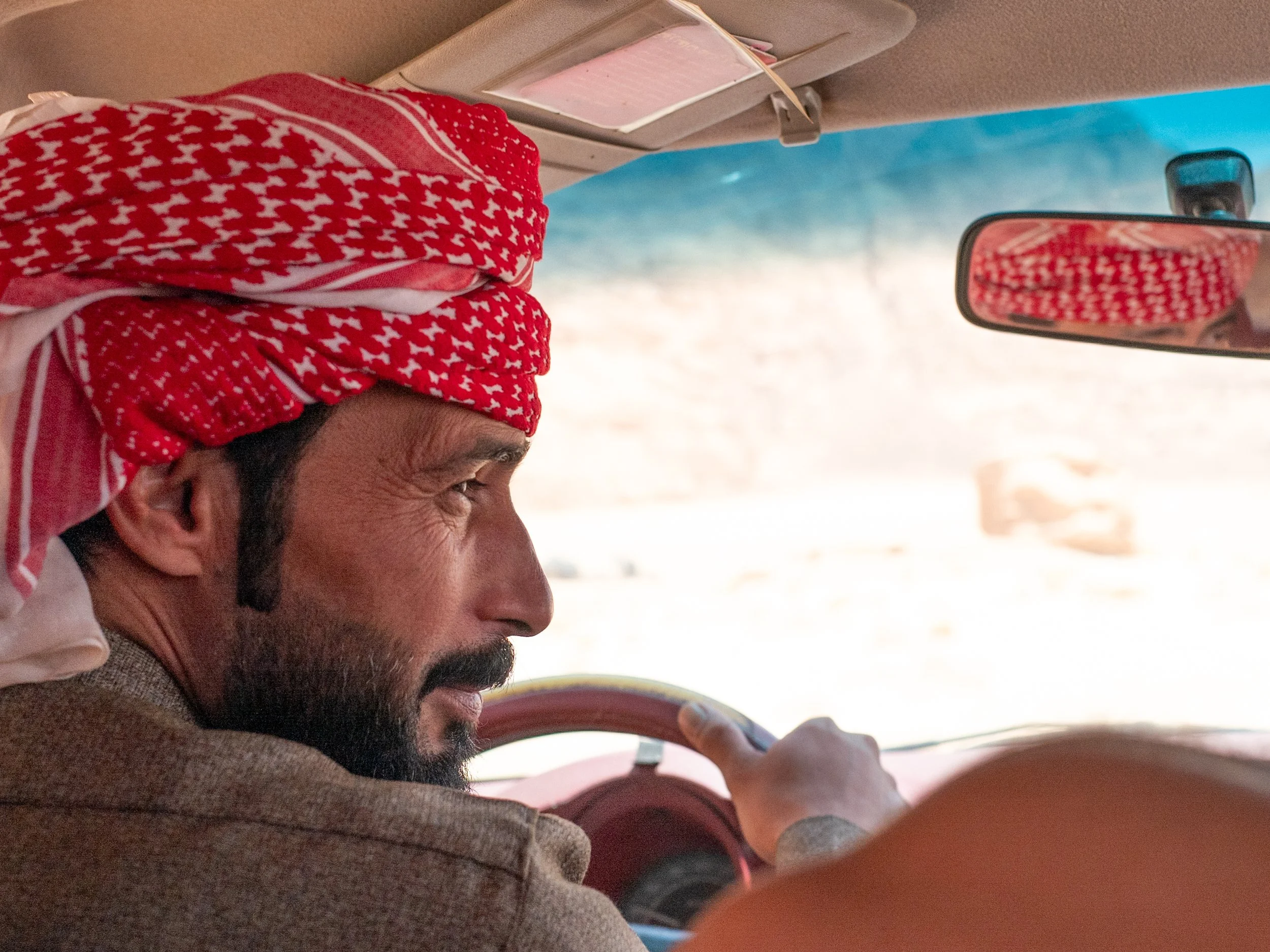 Close-up of a man with a red and white keffiyeh headscarf driving a vehicle in a desert landscape.