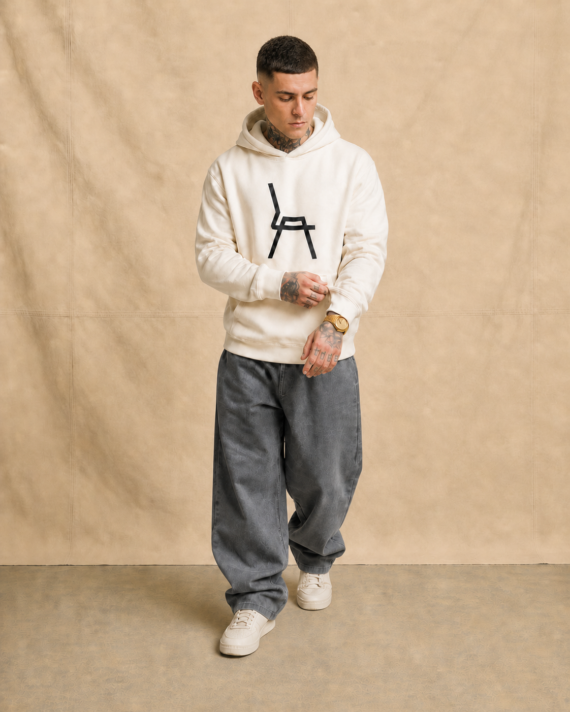 A young man with tattoos on his hands and neck, wearing a white hoodie with a black chair graphic, loose gray pants, white sneakers, and a gold watch, looking down and adjusting his sleeve against a beige background.