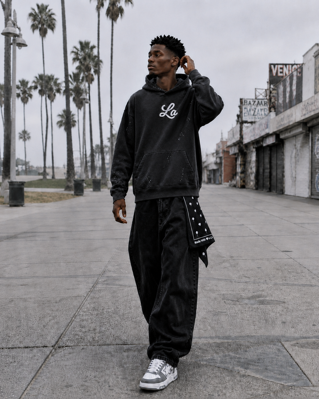 A young man standing on a deserted urban street with palm trees in the background, wearing a black hoodie, black pants, and sneakers.