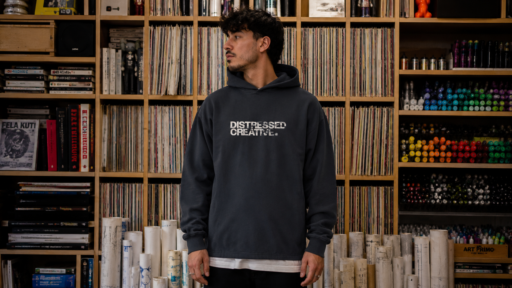 A young man with dark, curly hair and facial hair wearing a black hoodie that says 'Distressed Creative' standing in front of a wall filled with books, magazines, and art supplies.