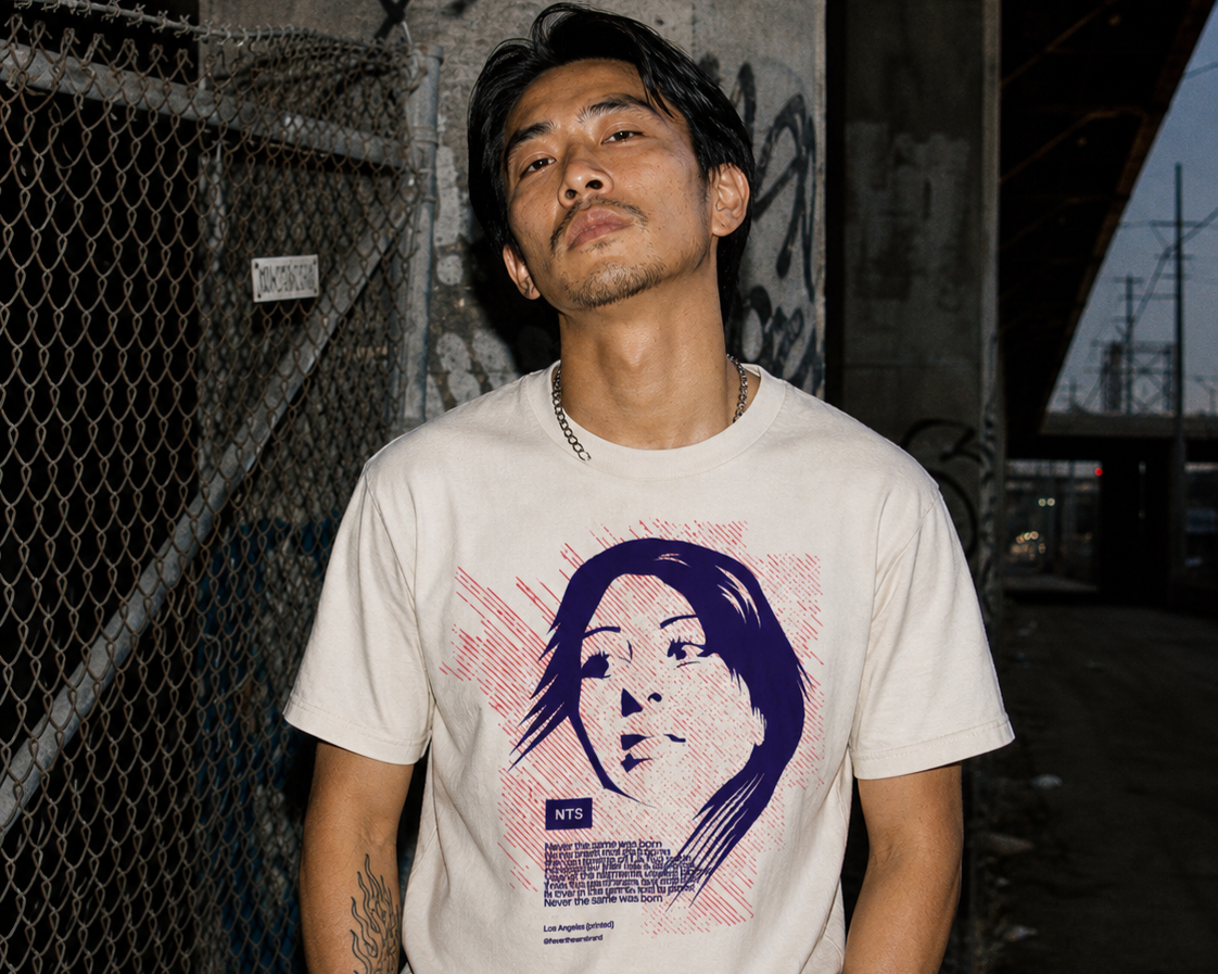 A man with black hair and a mustache stands in front of a chain-link fence and graffiti-covered wall. He is wearing a white T-shirt with a graphic of a woman's face and the text "NTS" and other smaller print. It appears to be evening or dusk.