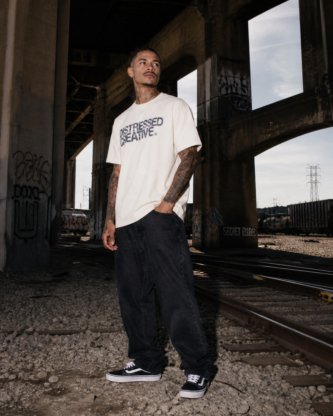 Man with tattoos in casual attire standing under a bridge near train tracks with graffiti, overcast sky in background.