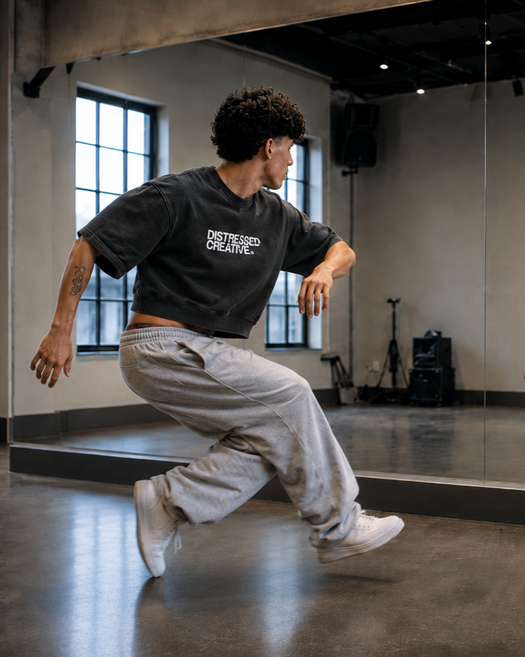 Person dancing in a studio with large windows and a mirror, wearing a black t-shirt and gray sweatpants.