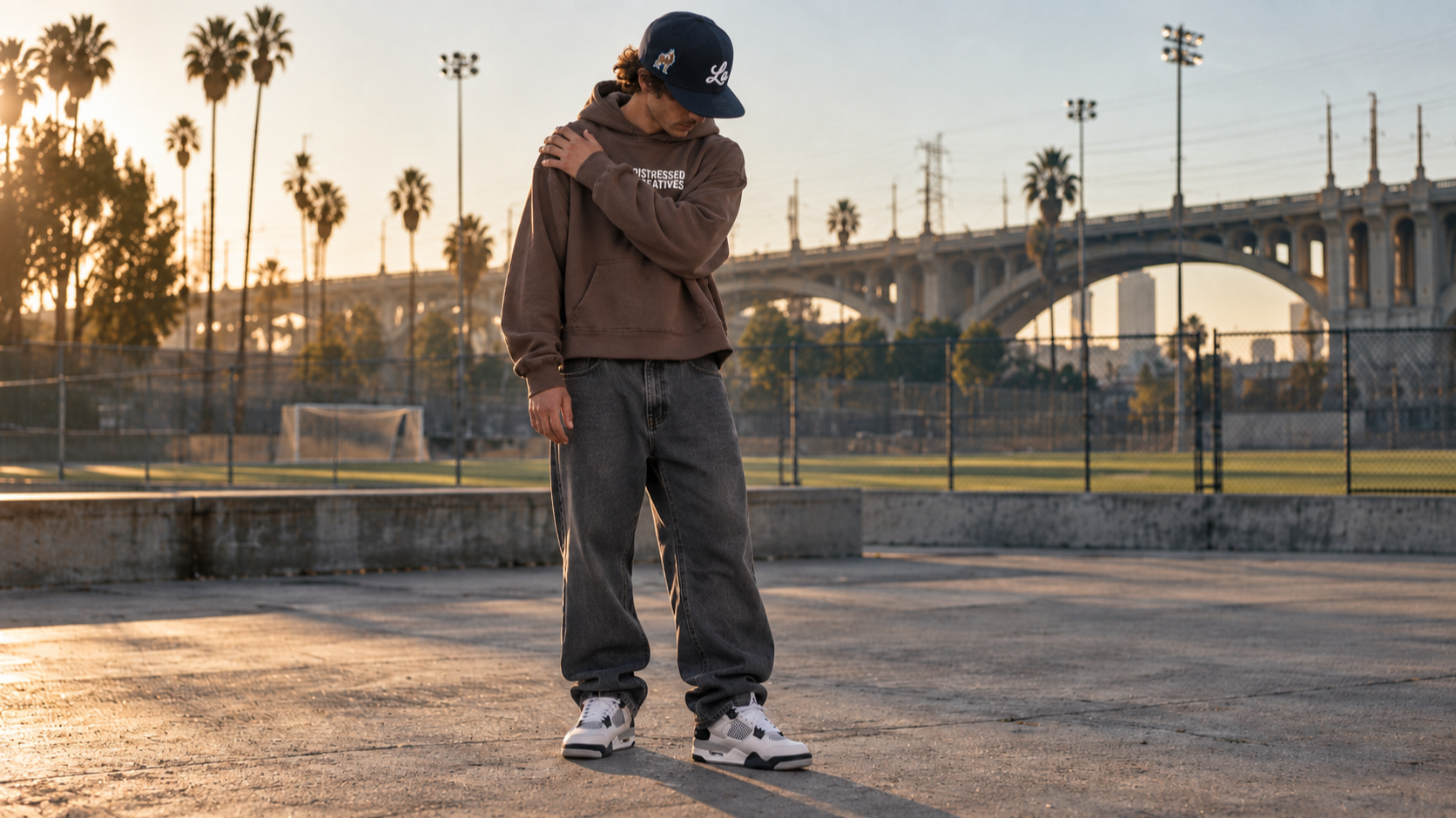 A young man standing on a skate park at sunset, wearing a brown La Visual Studios hoodie, loose jeans, sneakers, and a La Visual Studios special edition baseball hat, with palm trees and a bridge in the background.
