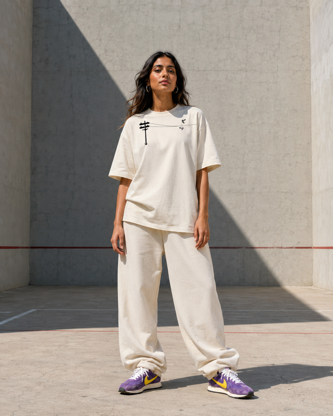 A woman standing outdoors on a concrete surface against a light gray wall, wearing an oversized beige T-shirt with black graphic print, wide beige pants, and purple sneakers with yellow accents.