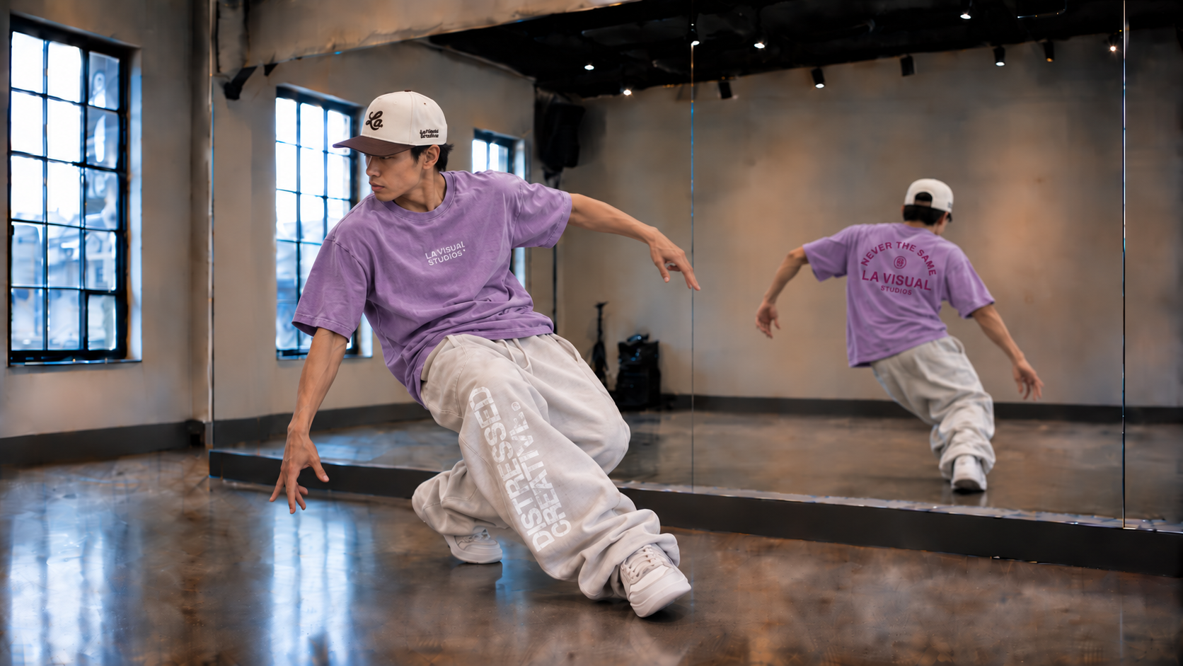 A young Asian male hip-hop dancer practicing in a dance studio wearing a La Visual Studios two-tone cream and brown hat and  purple La Visual Studios t-shirt and white baggy LVS sweatpants with his reflection visible in a mirror.