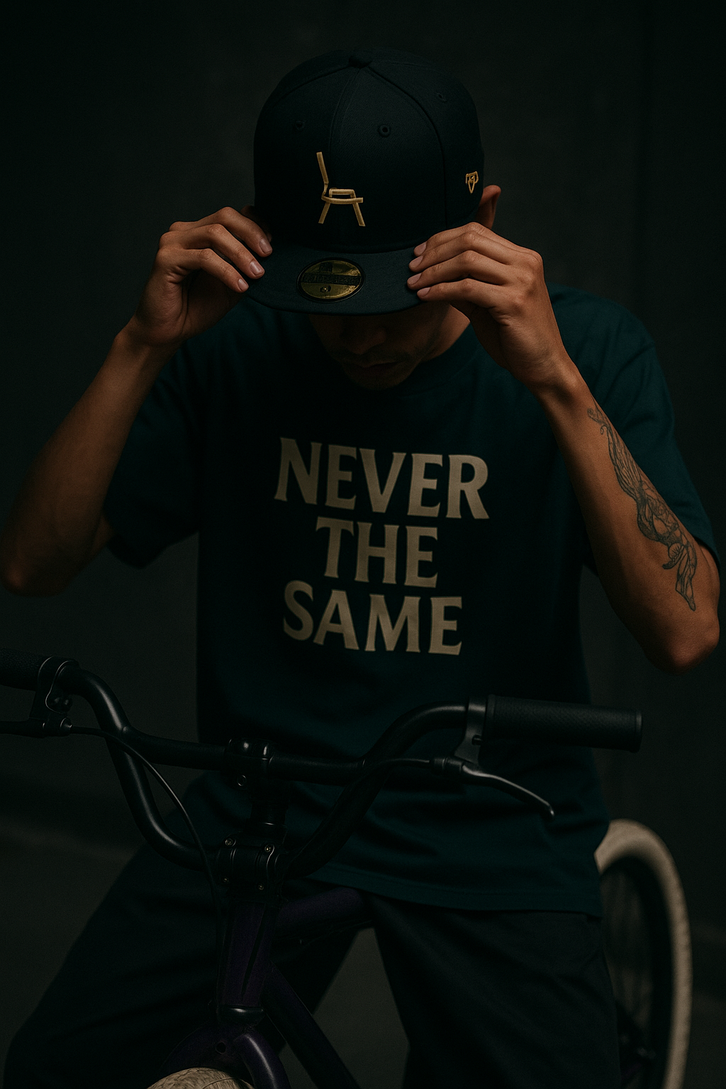 A person wearing a dark baseball cap and a t-shirt that says "NEVER THE SAME" while holding the bill of their cap. The person is sitting on a bicycle, and has a visible tattoo on their right arm.