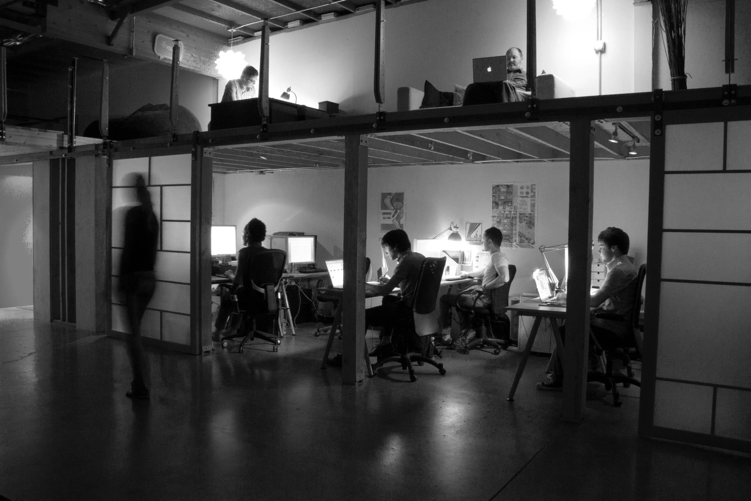 An office space with five people working at their desks, and two individuals on a raised loft area working on computers. The office has warm lighting, and there's a blurred person walking past.