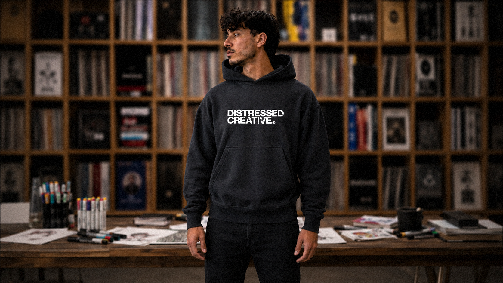 A young man with curly dark hair and light facial hair wearing a black hoodie with 'DISTRESSED CREATIVE' printed on it, standing in front of a bookshelf filled with books and artwork in a creative space.