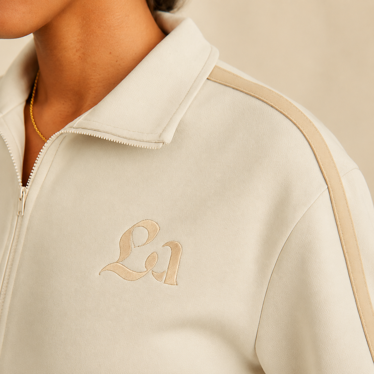Close-up of a person wearing a cream-colored jacket with the initials 'La' embroidered on the chest, zipper closure, and beige accents on the shoulder, with a gold necklace visible around the neck.