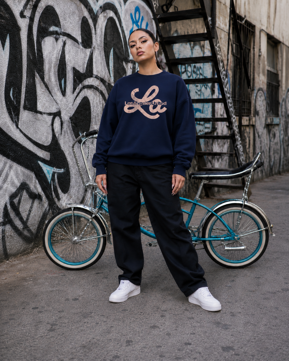 A woman standing in front of a wall with graffiti, holding a bicycle, wearing a dark blue Los Angeles sweatshirt, black pants, and white sneakers.
