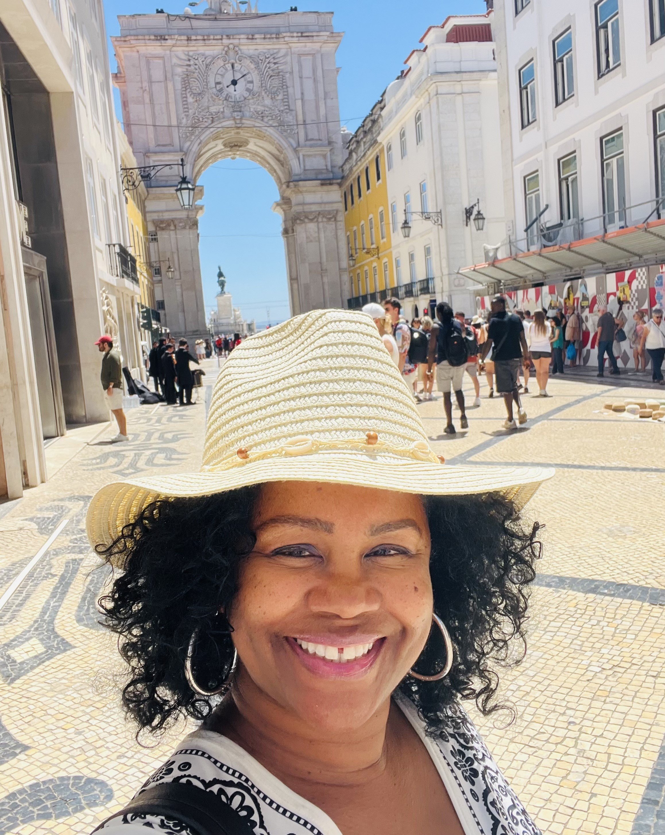 Smiling woman wearing a wide-brimmed hat and hoop earrings takes a selfie in a pedestrian street with historic architecture and a large archway in Lisbon, Portugal, on a sunny day.