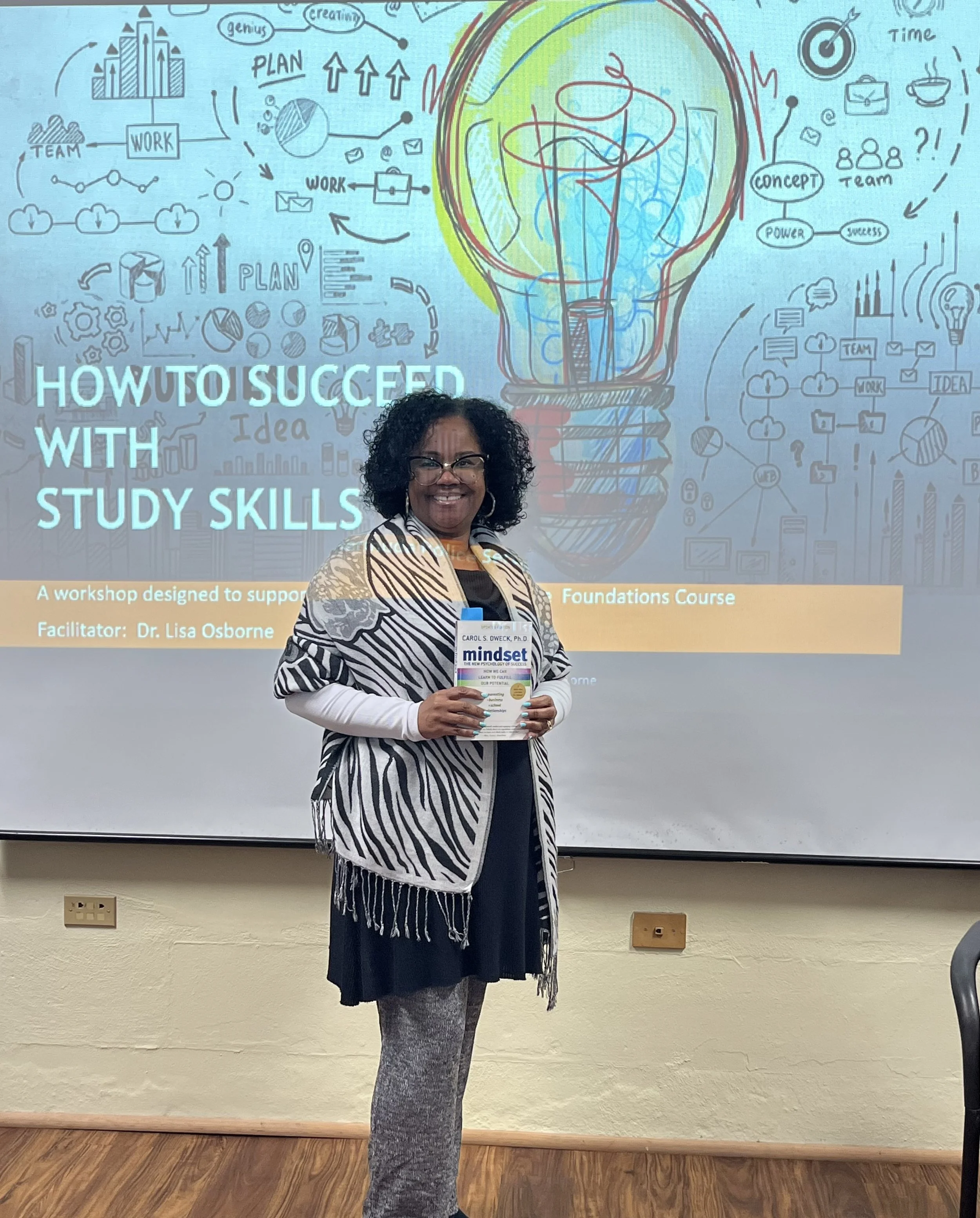A woman standing in front of a presentation slide titled 'How to Succeed with Study Skills'. She is holding a book titled 'Mindset'. The background slide shows a large illustrated light bulb and various graphs, charts, and icons related to planning, teamwork, and ideas.