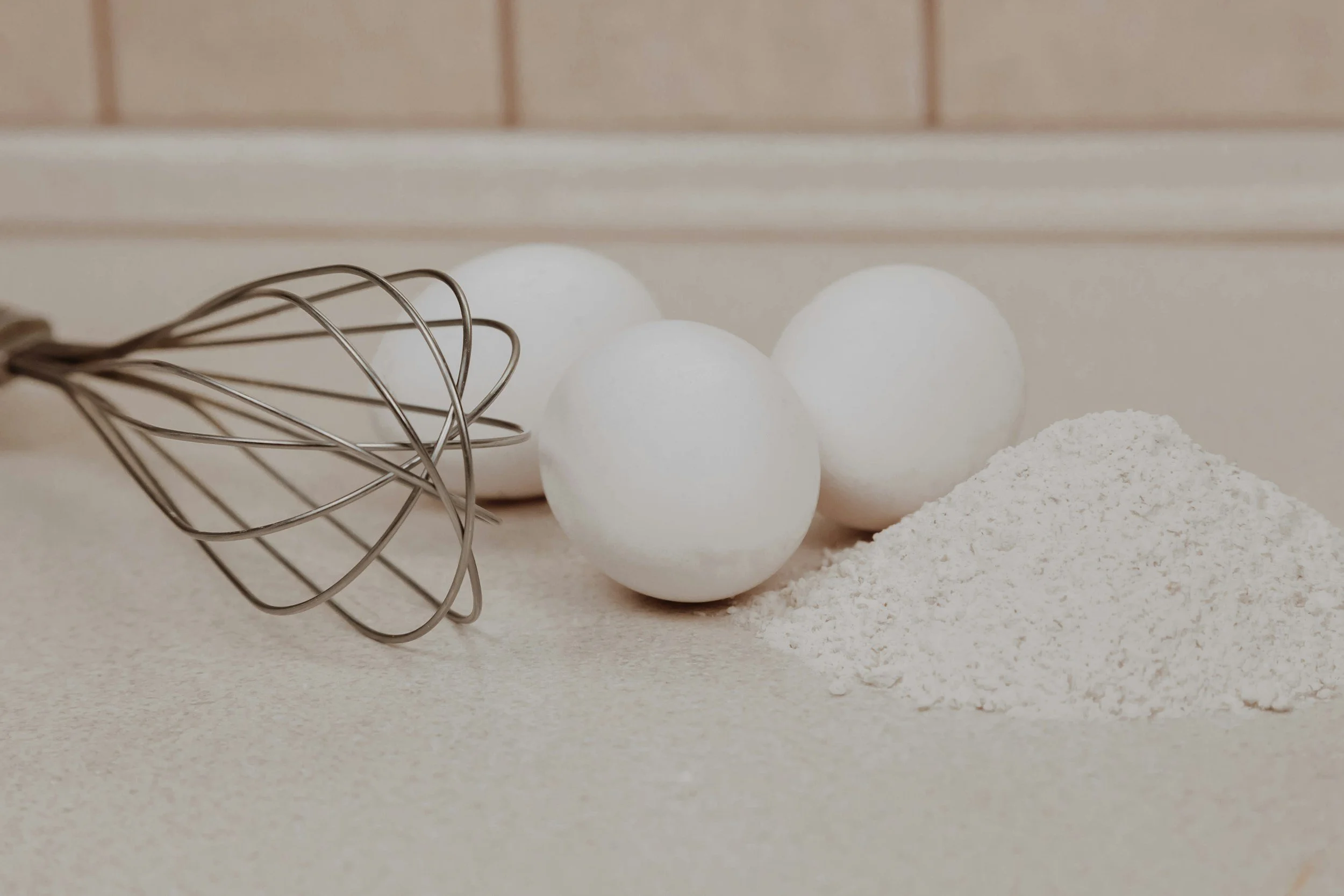 Three white eggs, a pile of flour, and a metal whisk on a kitchen countertop.