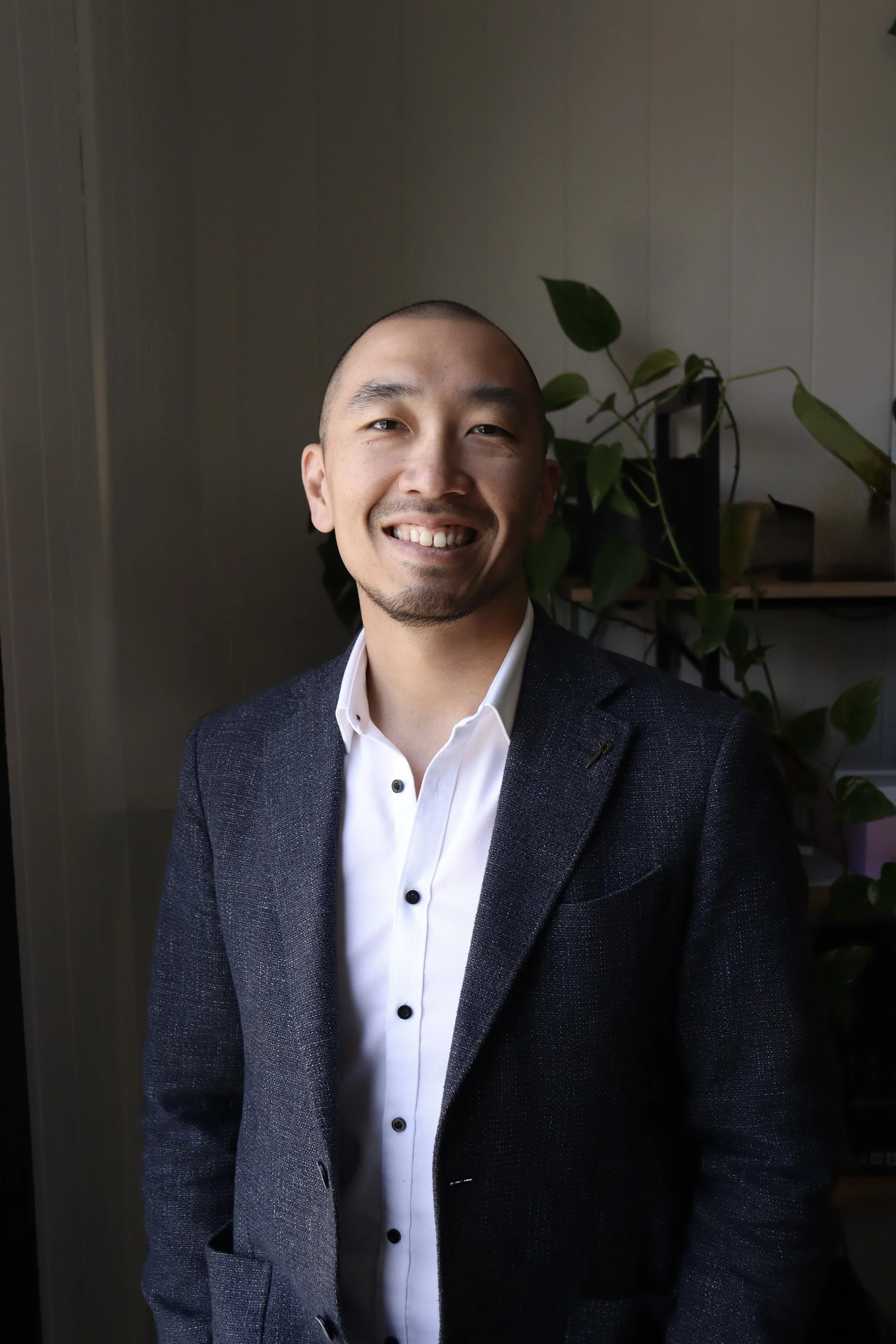 Smiling man with a shaved head in a dark blazer and white shirt standing indoors near a window with plants in the background.