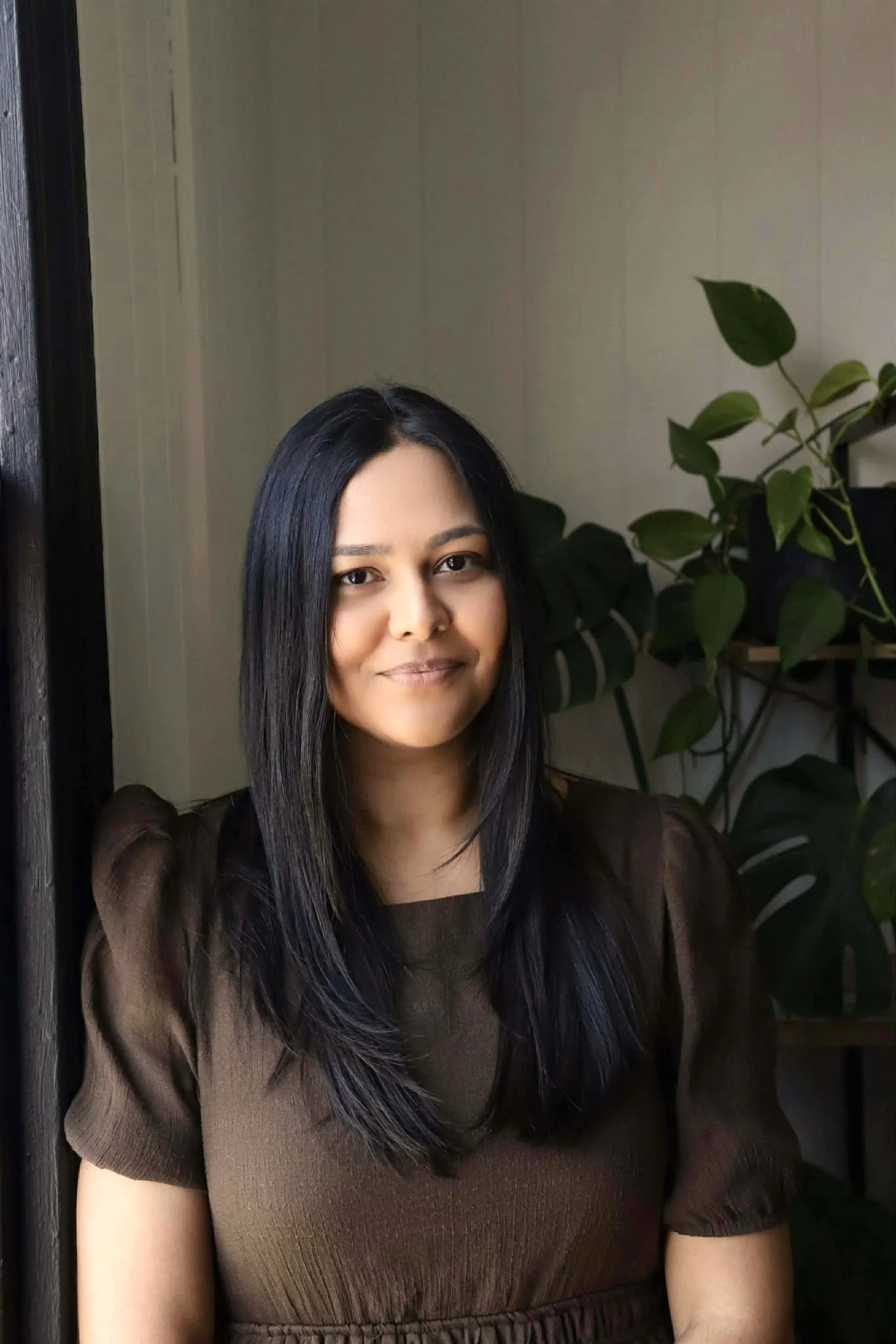 A woman with long black hair in a brown top, sitting indoors near a window with potted plants in the background.