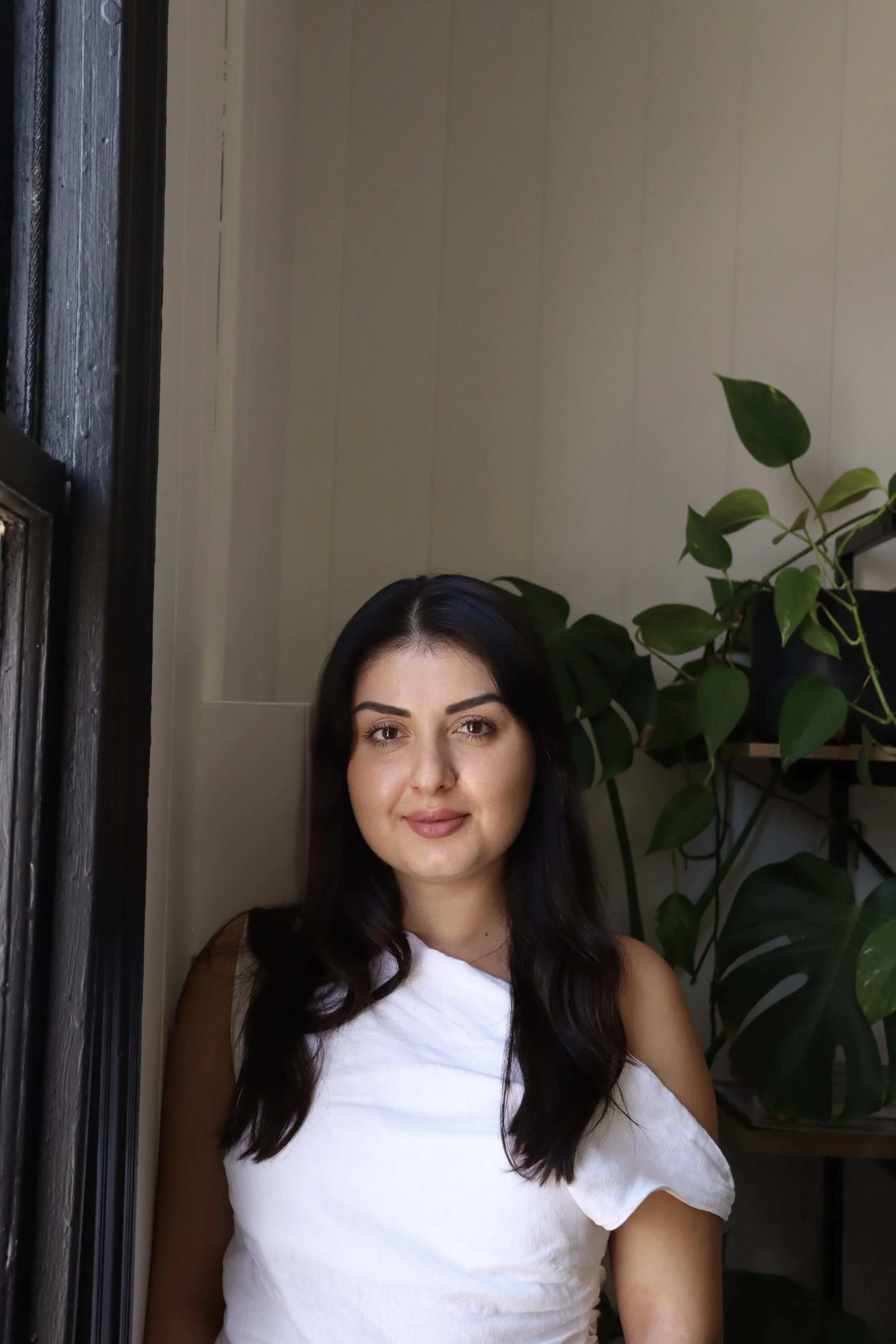 A woman with dark hair, wearing a white top, standing indoors near green plants and a window with dark trim.
