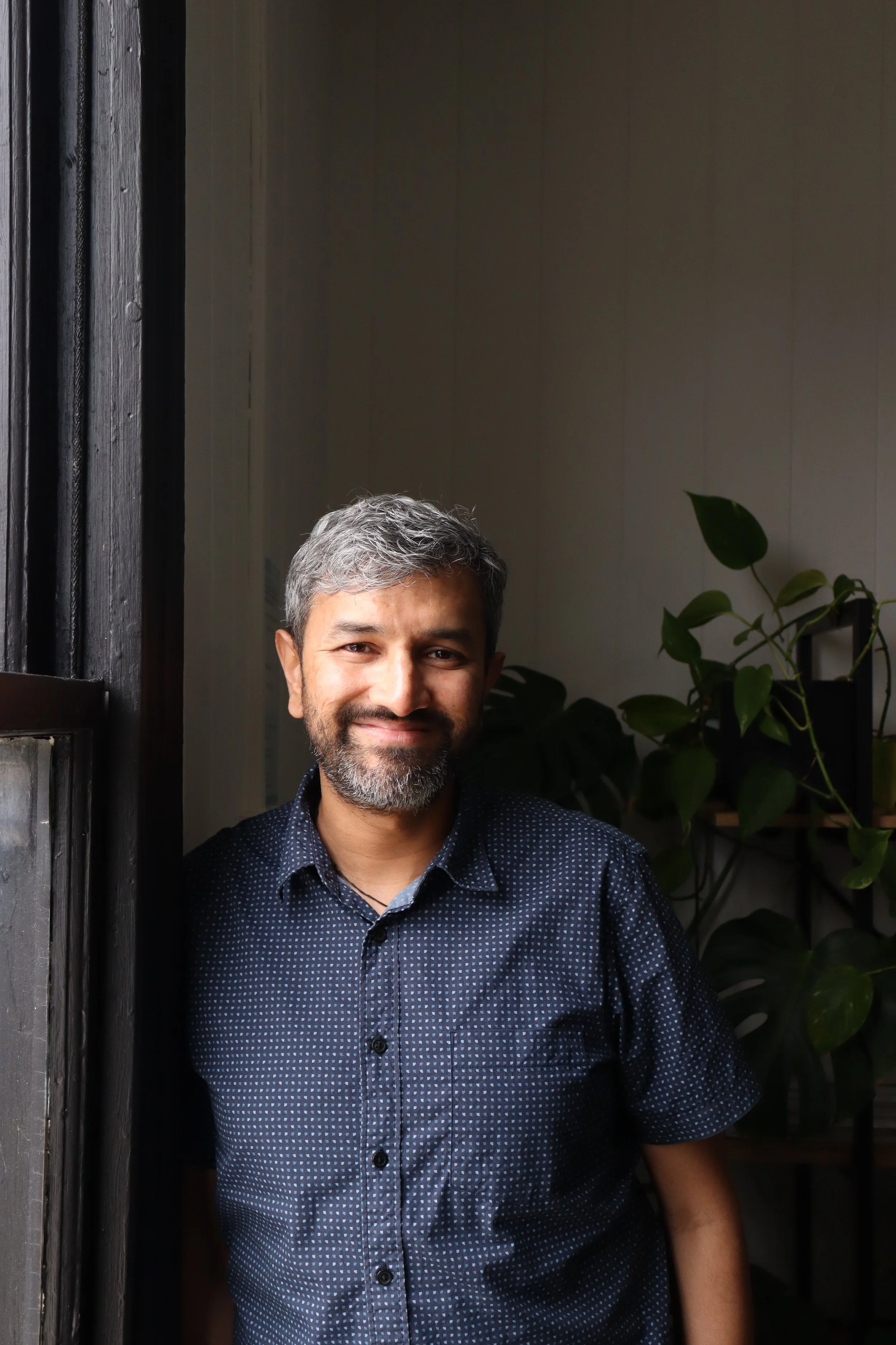 Man with gray hair and beard smiling indoors near window, with green plants in background.