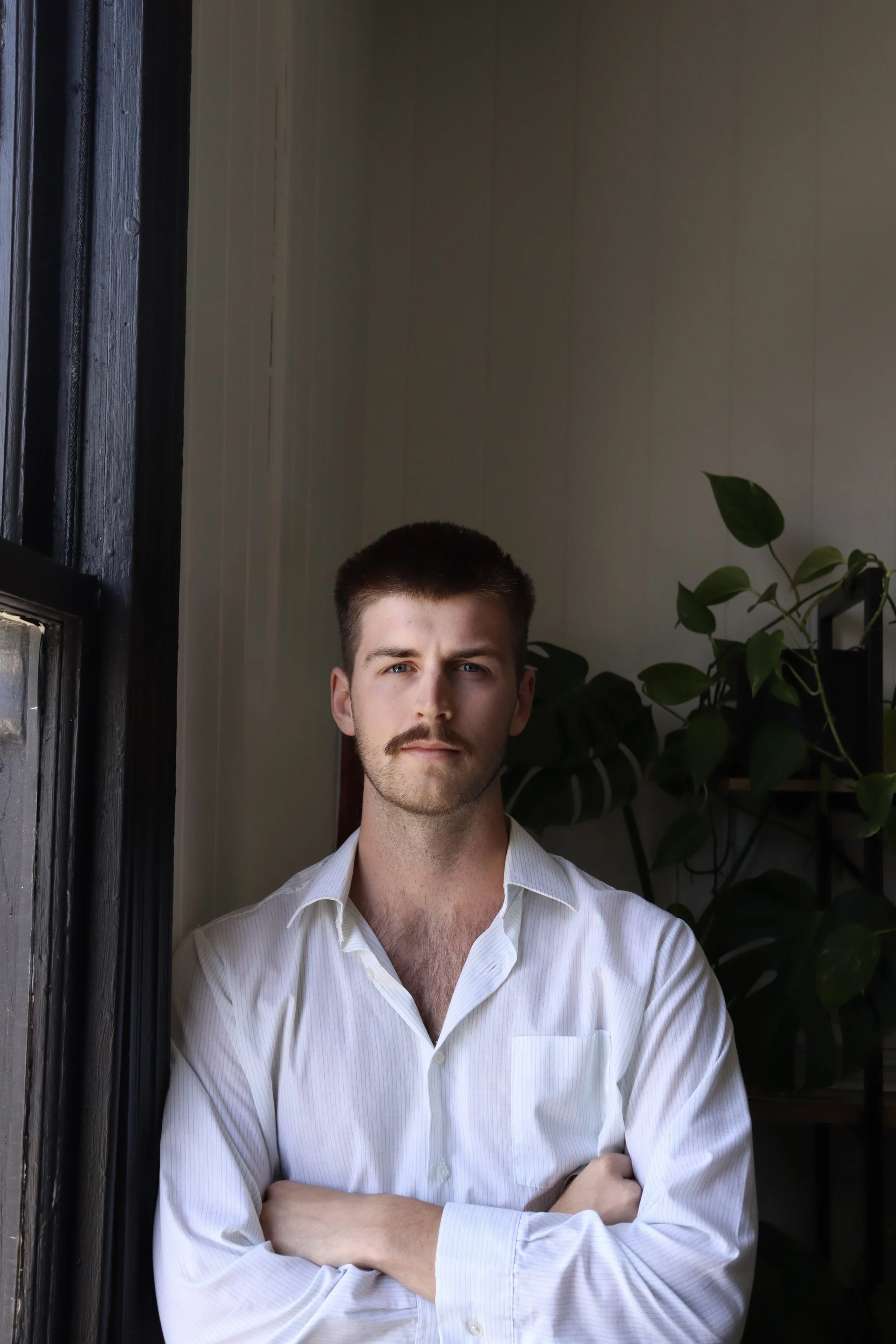 A man with short brown hair, a mustache, and a beard, standing indoors next to a window, wearing a white dress shirt with his arms crossed, with green plants and shelves in the background.