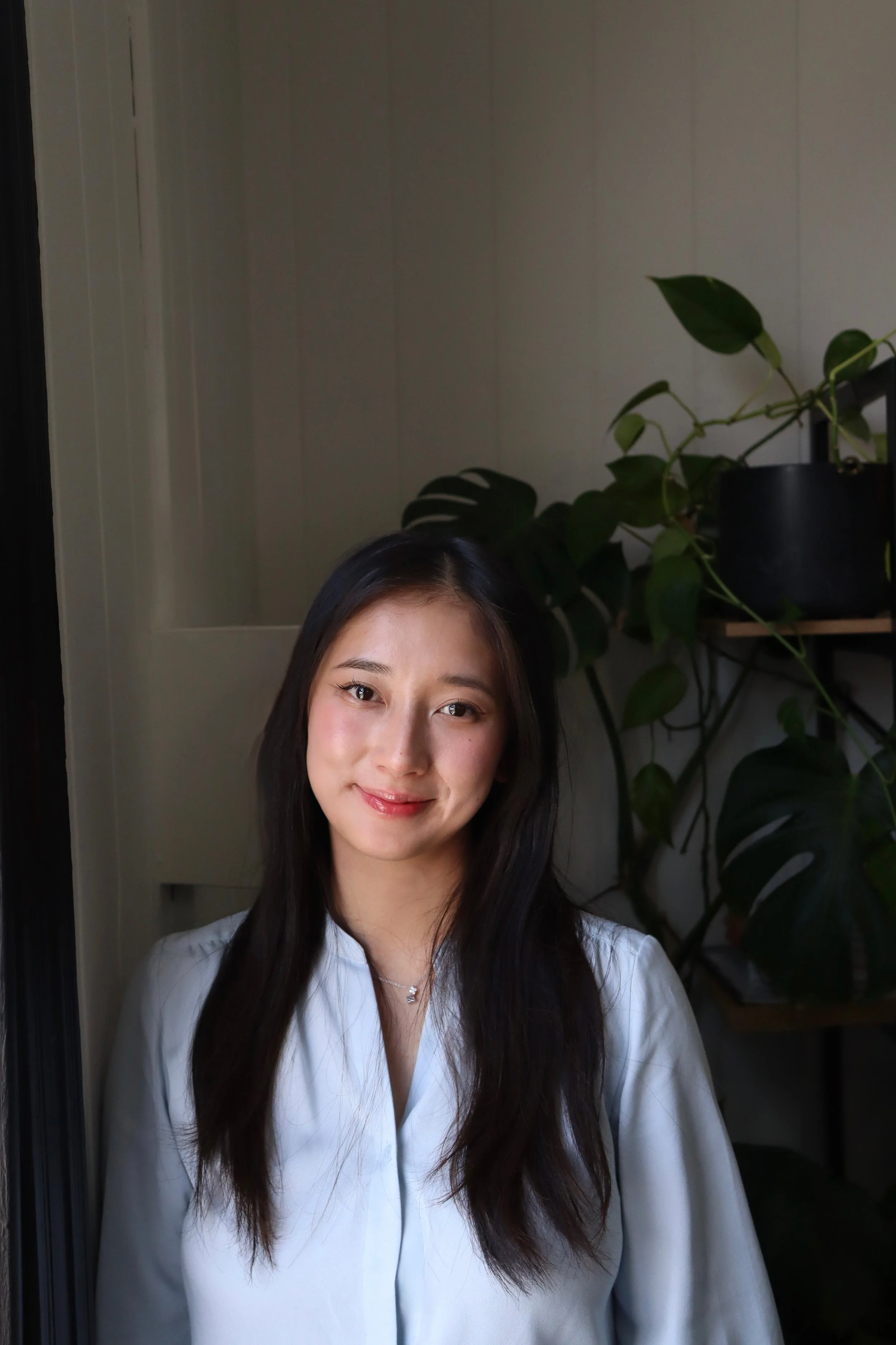 A young woman with long dark hair smiling, wearing a white blouse, standing indoors next to a potted plant.