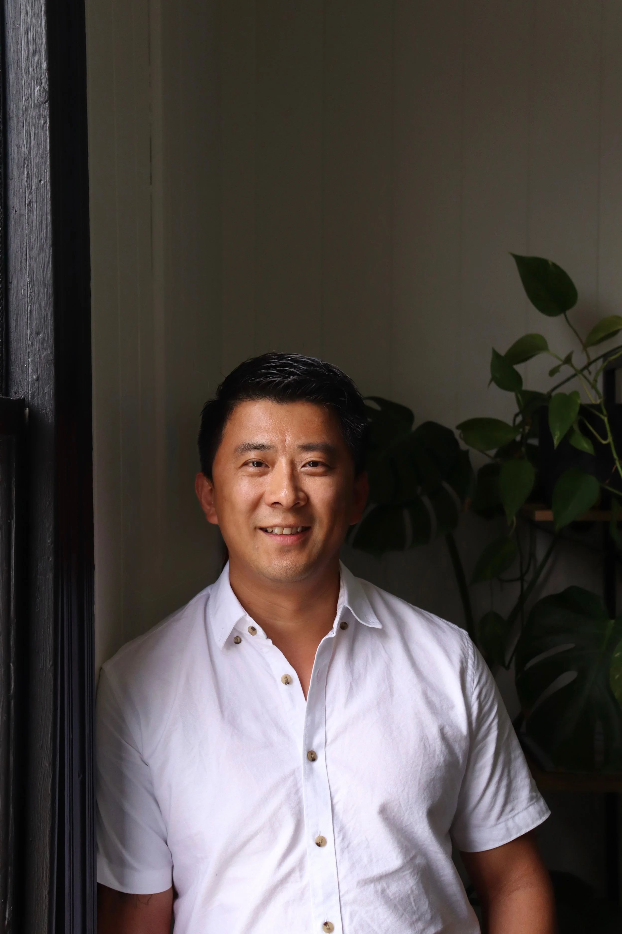 A man smiling, wearing a white button-up shirt, standing in front of a dark wooden door frame with indoor plants in the background.