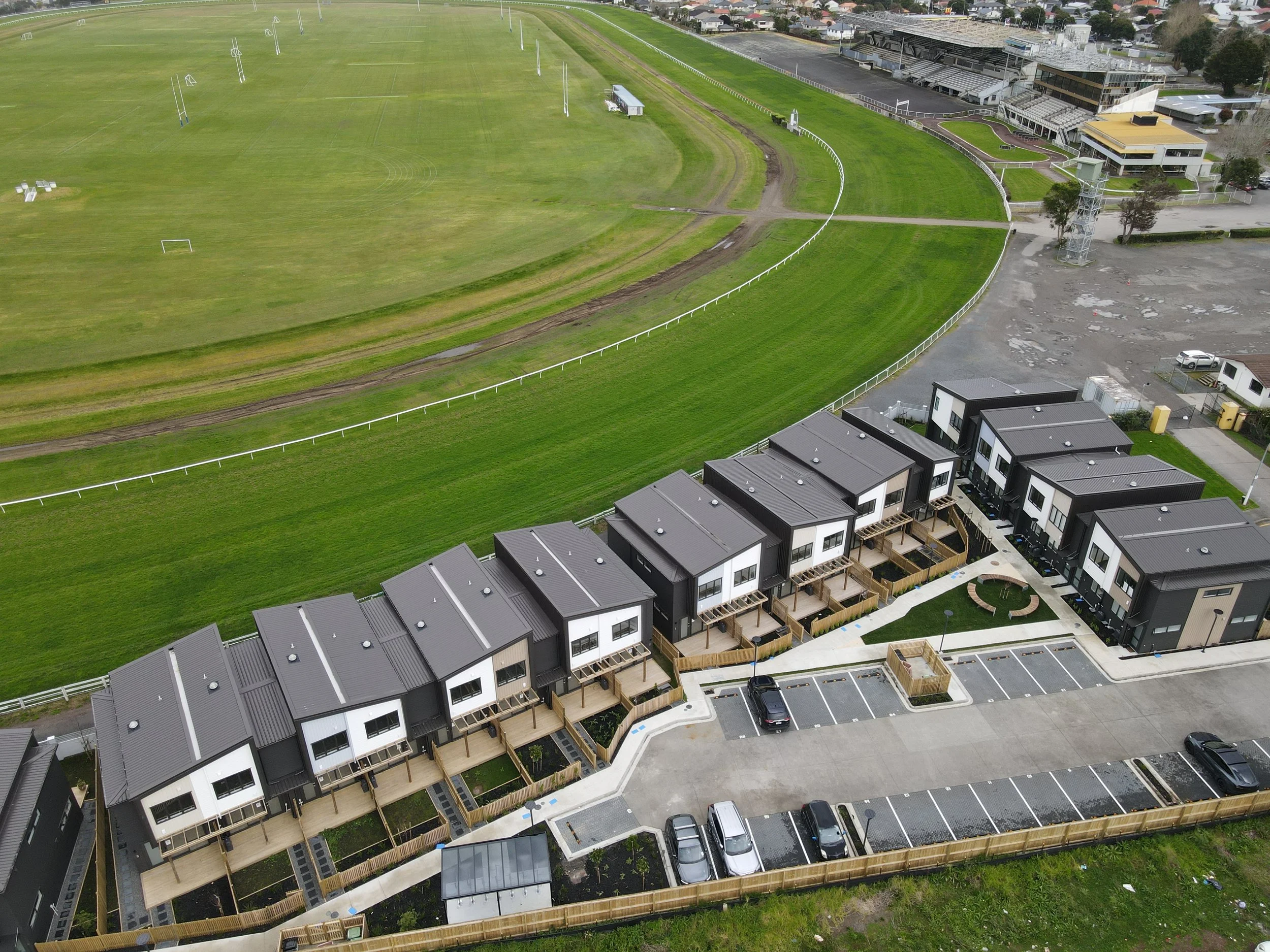 An aerial view of a row of modern townhouses with black and white facades, small decks, and parking lots in front, next to a large green horse racing track with white fencing.