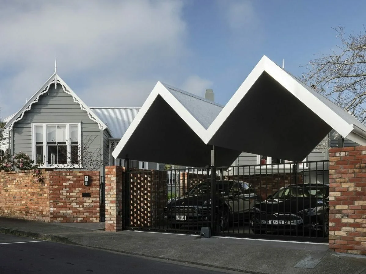 A modern house with a traditional gabled house in the background, a brick wall, and a gate with a black and white geometric overhang, with cars parked inside.