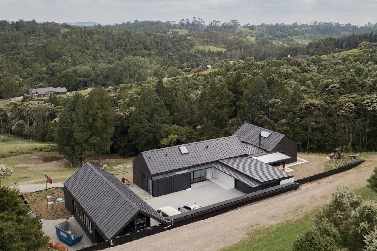 Aerial view of a modern black metal roof house in a lush green rural area with trees and rolling hills.