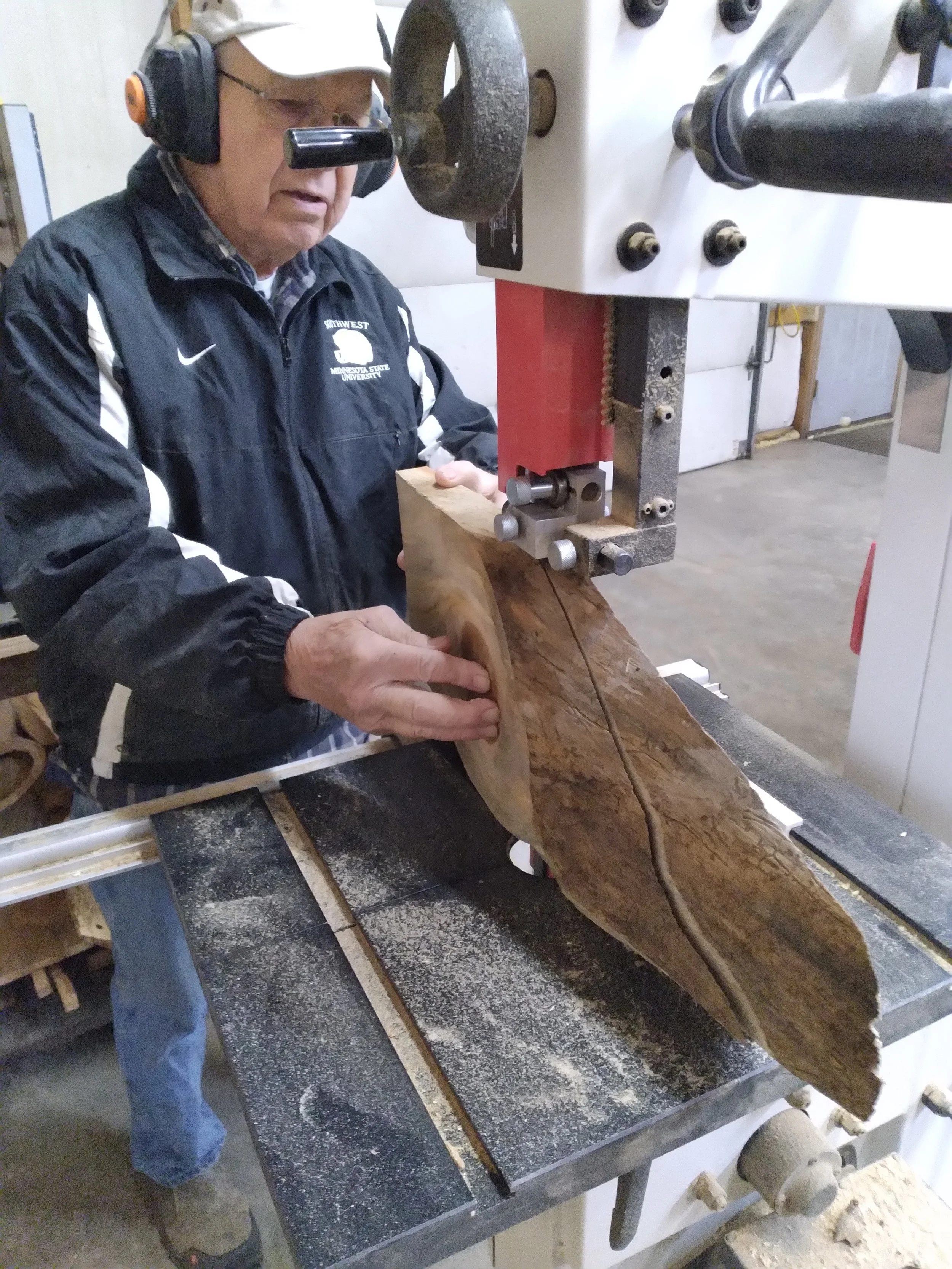 A man in safety gear operating a bandsaw to cut a large piece of wood in a workshop.