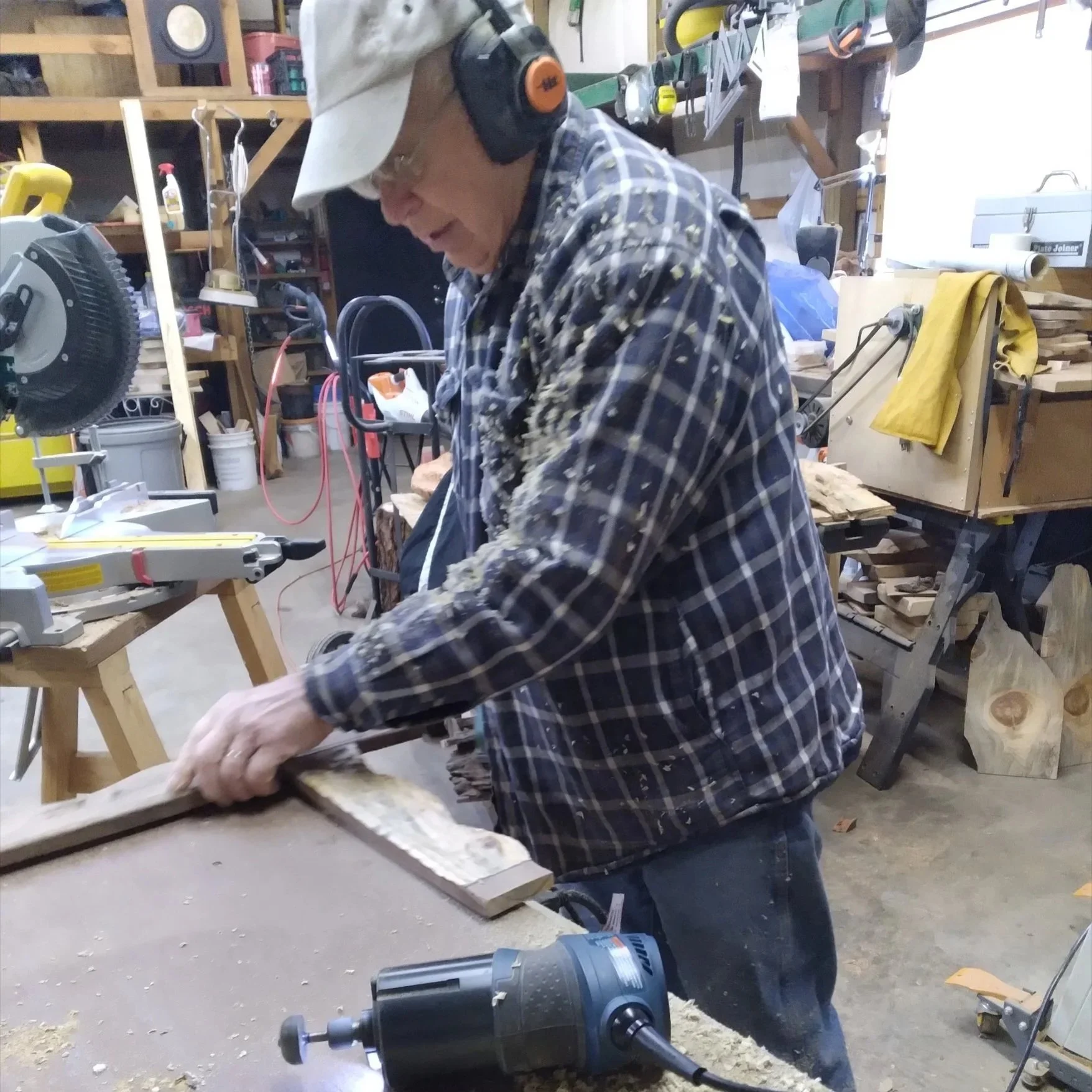 An elderly man in a plaid shirt and baseball cap working in a woodworking shop. He is wearing ear protection and is sanding a wooden piece on a workbench covered in sawdust, with various woodworking tools and equipment in the background.