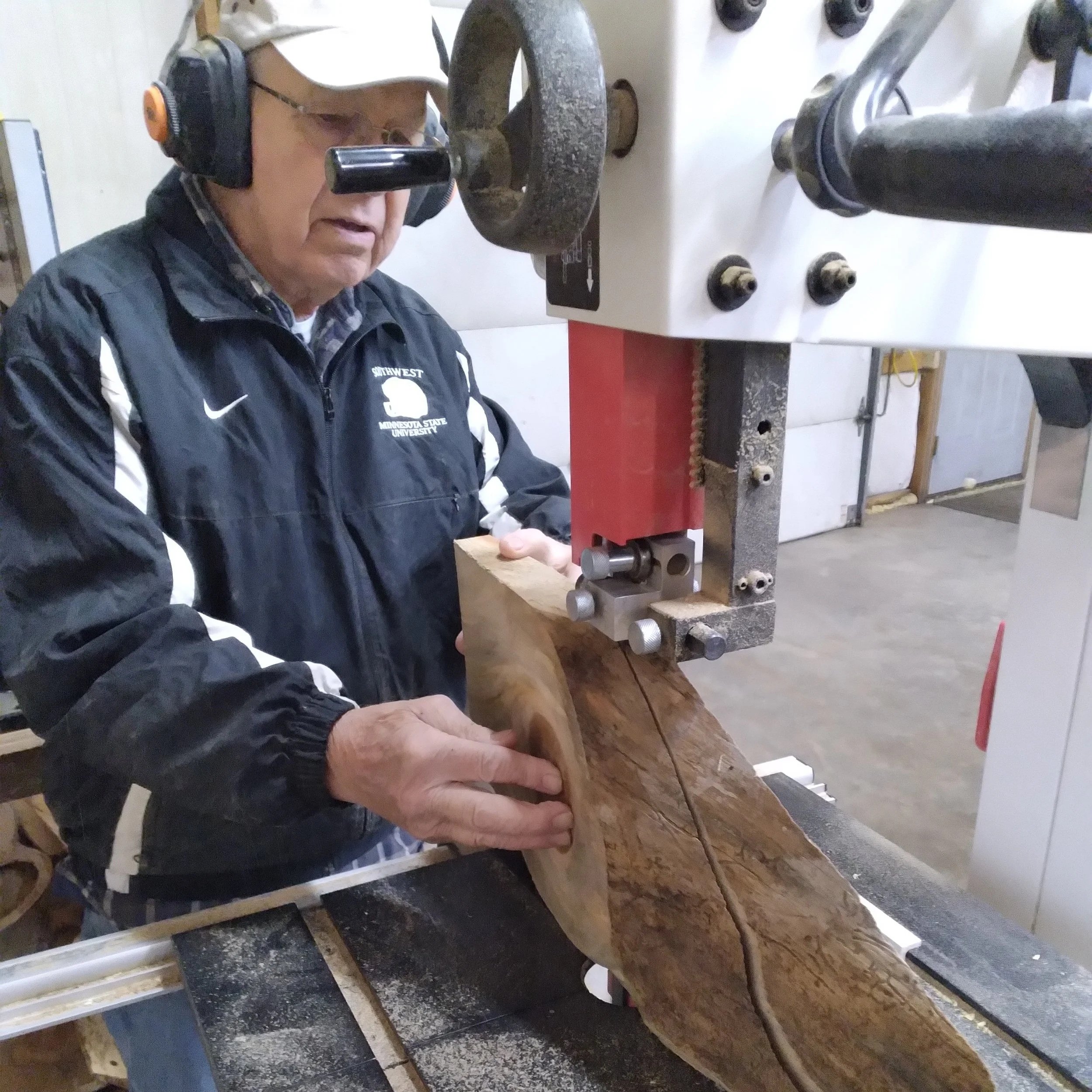 An elderly man with glasses, a baseball cap, and noise-canceling headphones is operating a bandsaw to cut a large piece of wood in a workshop. He is wearing a black jacket with white stripes and a logo, and is guiding the wood through the saw.