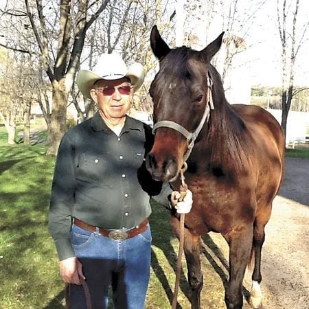 An older man wearing a cowboy hat, sunglasses, and a black shirt stands next to a brown horse with a white stripe on its face outdoors in a park.