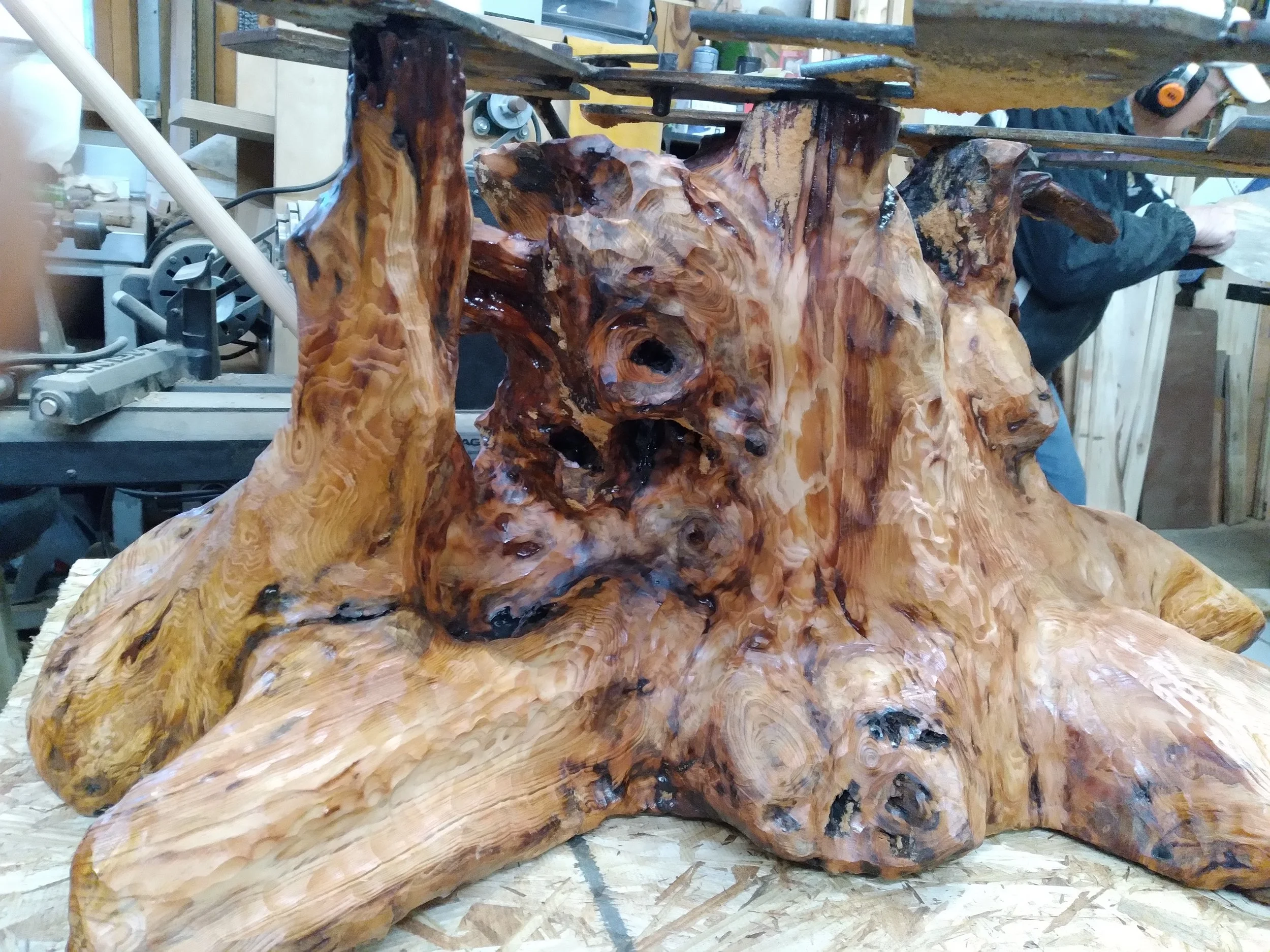 A large, polished wood sculpture with an abstract, organic shape and natural wood grain patterns, located in a woodworking workshop.