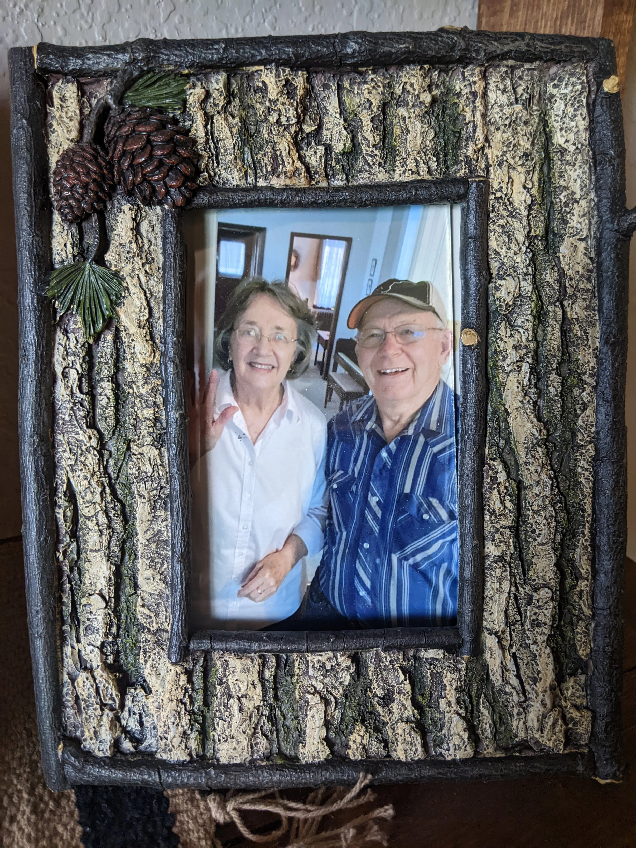 A framed photo of an elderly couple smiling and posing indoors, with a rustic, bark-textured wooden frame decorated with pinecones and pine leaves.