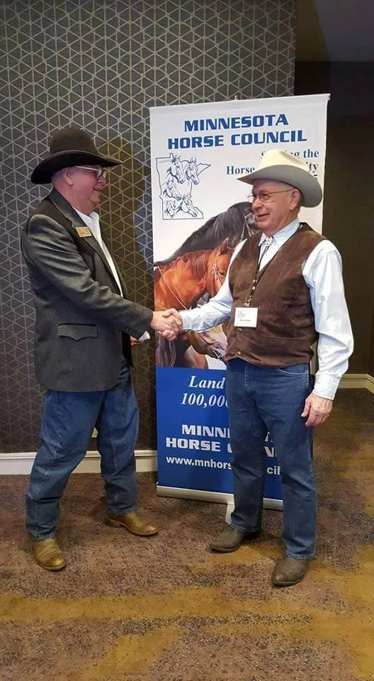 Two men in cowboy hats shaking hands at a Minnesota Horse Council event, standing in front of a banner with images of horses and the council's name.