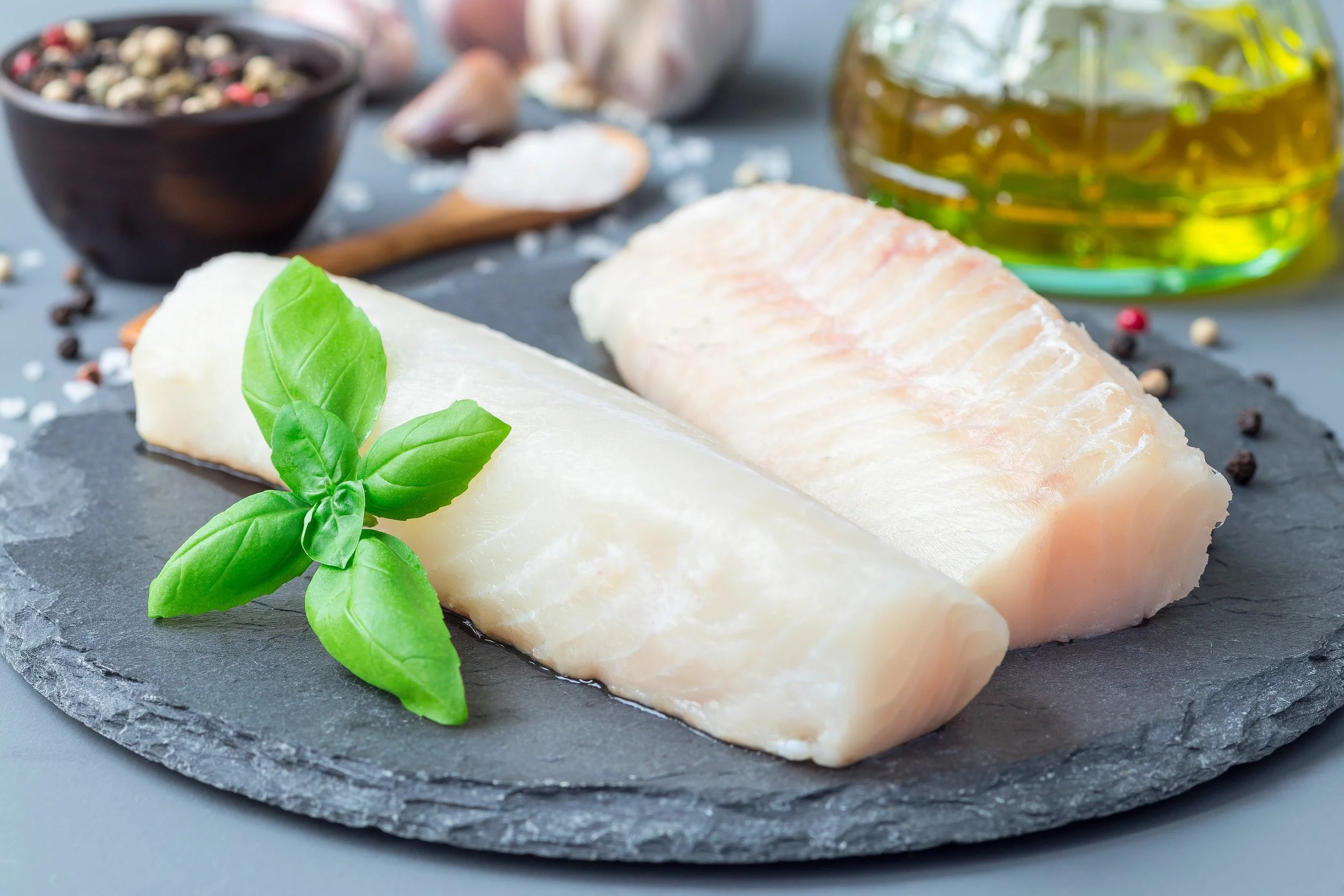 Fresh raw fish fillets with basil leaves on a black slate platter, with salt, peppercorns, and oil in the background.