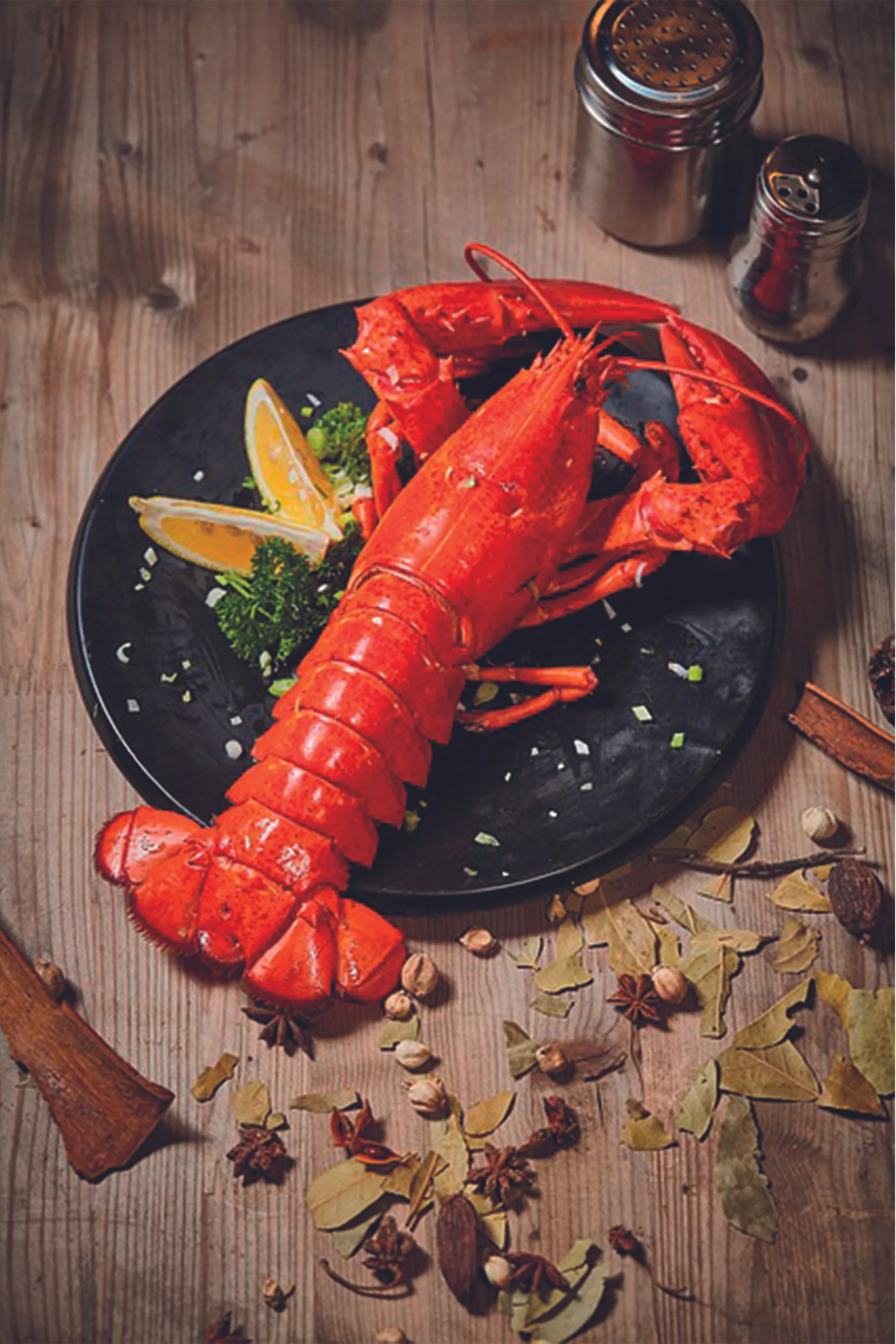 A cooked lobster on a black plate garnished with lemon wedges, parsley, and chopped green onions, placed on a wooden table with scattered bay leaves, cinnamon sticks, star anise, and peppercorns, next to salt and pepper shakers.