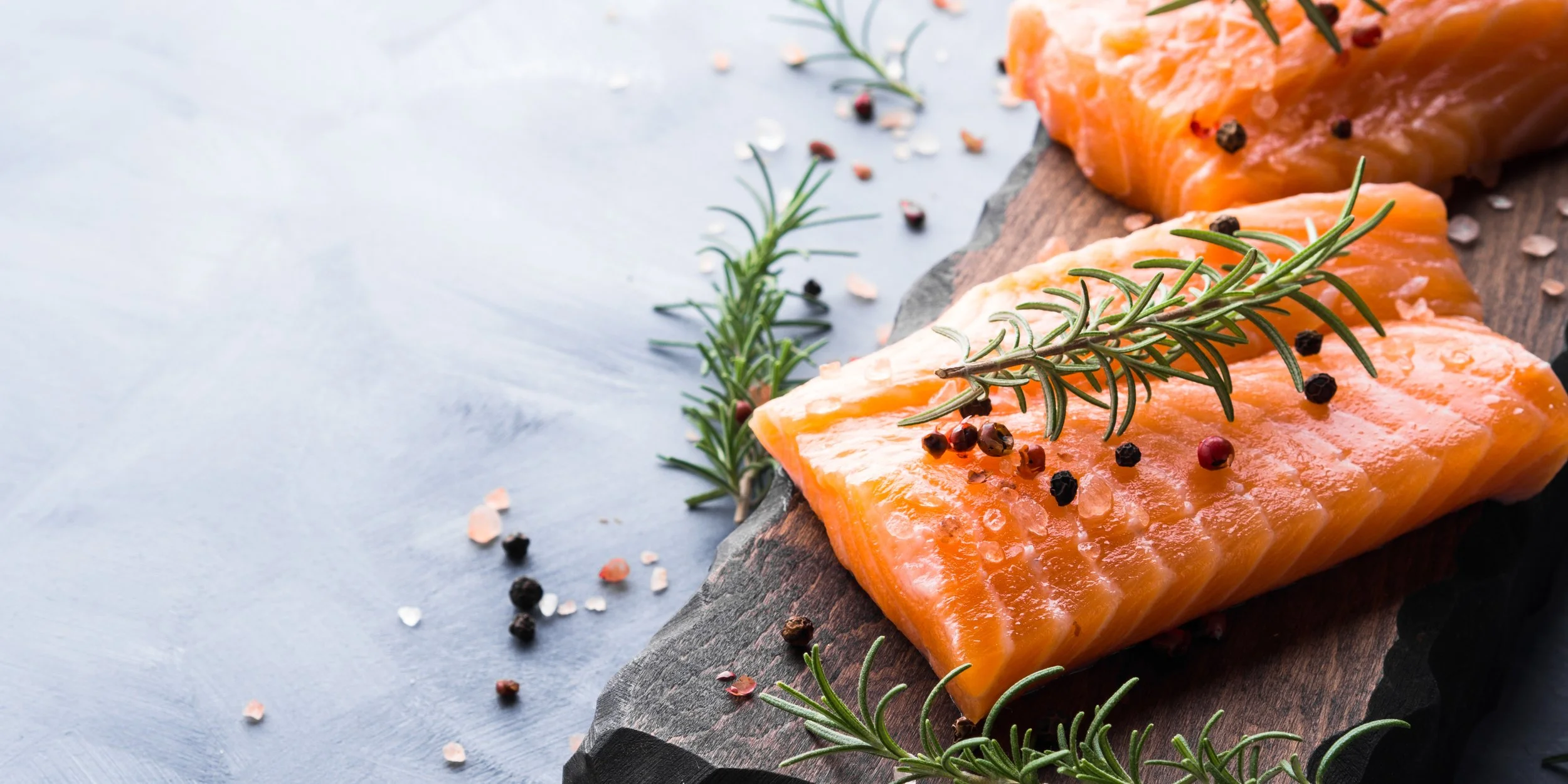 Fresh raw salmon fillets garnished with rosemary sprigs and black and pink peppercorns on a dark wooden board