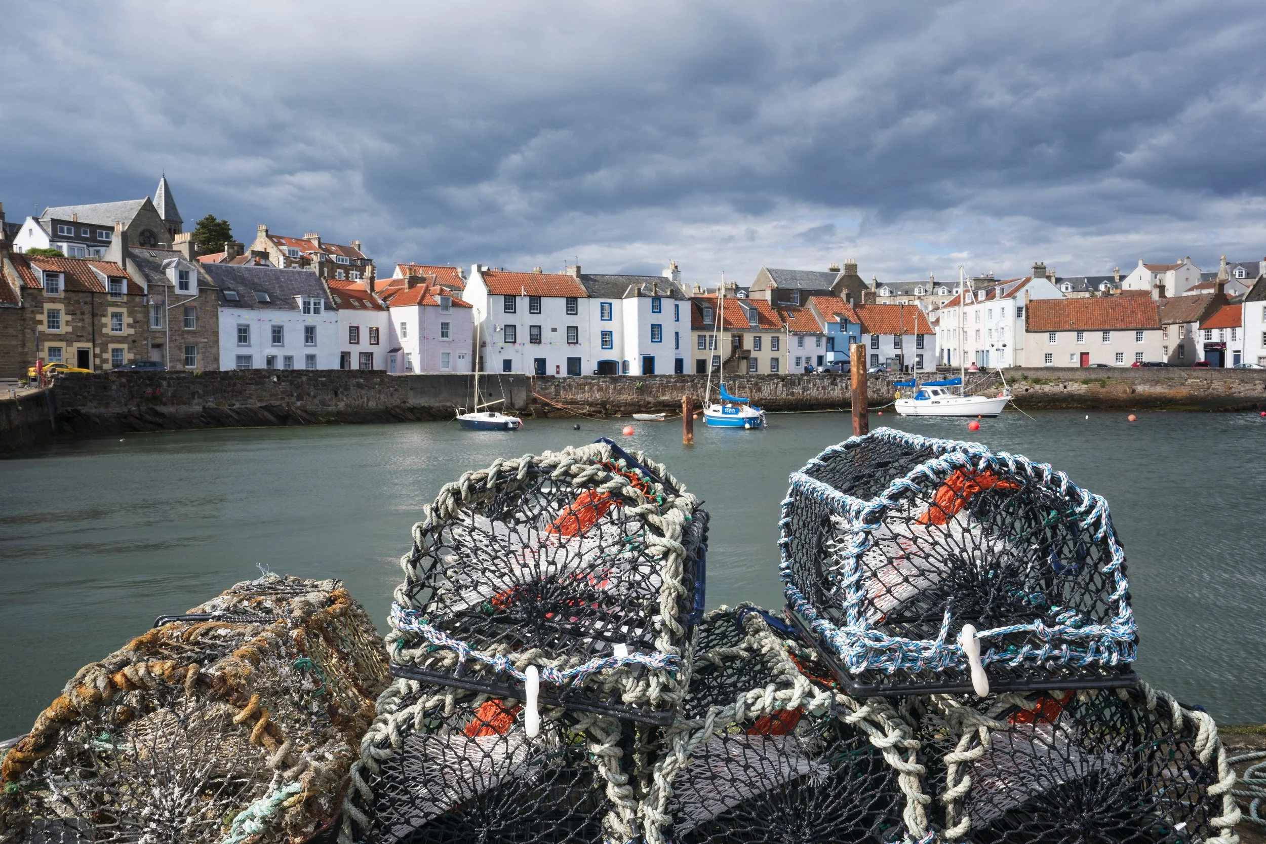Lobster traps stacked on a dock with a view of colorful houses and boats in a harbor under a cloudy sky.