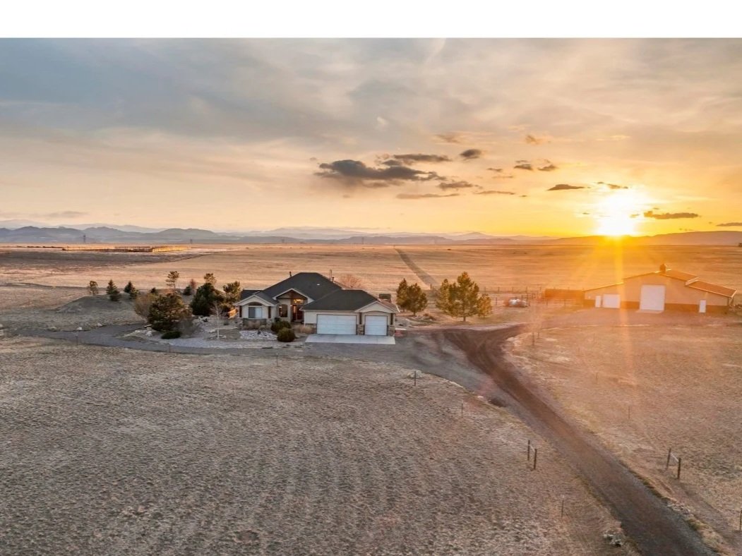 A rural property with a house and trees on a flat, open landscape at sunset, with a dirt driveway and distant mountains in the background.