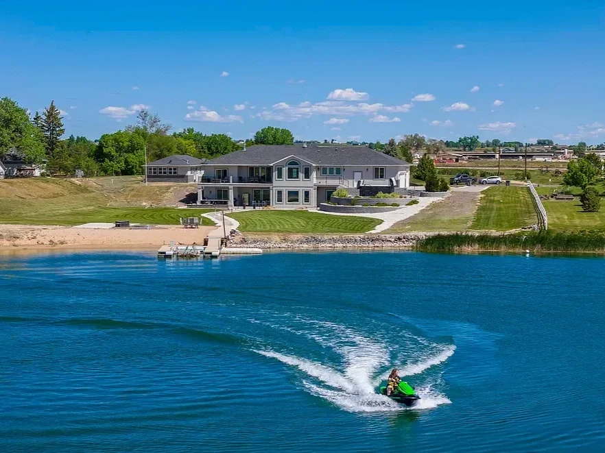 A person riding a jet ski on a lake with a large house and green lawn in the background under a blue sky with scattered clouds.