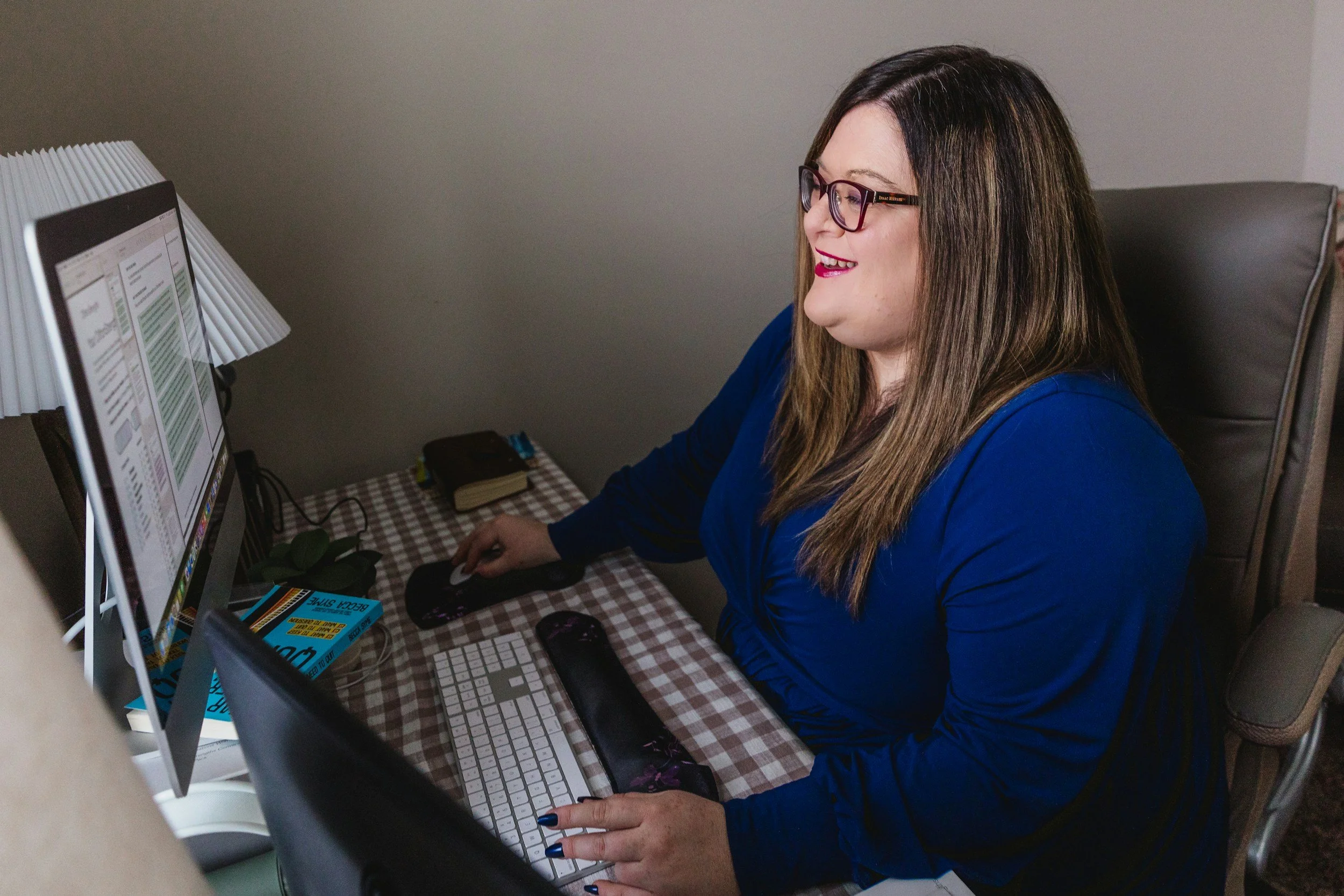 Woman in glasses smiling while working at a desktop computer in a home office