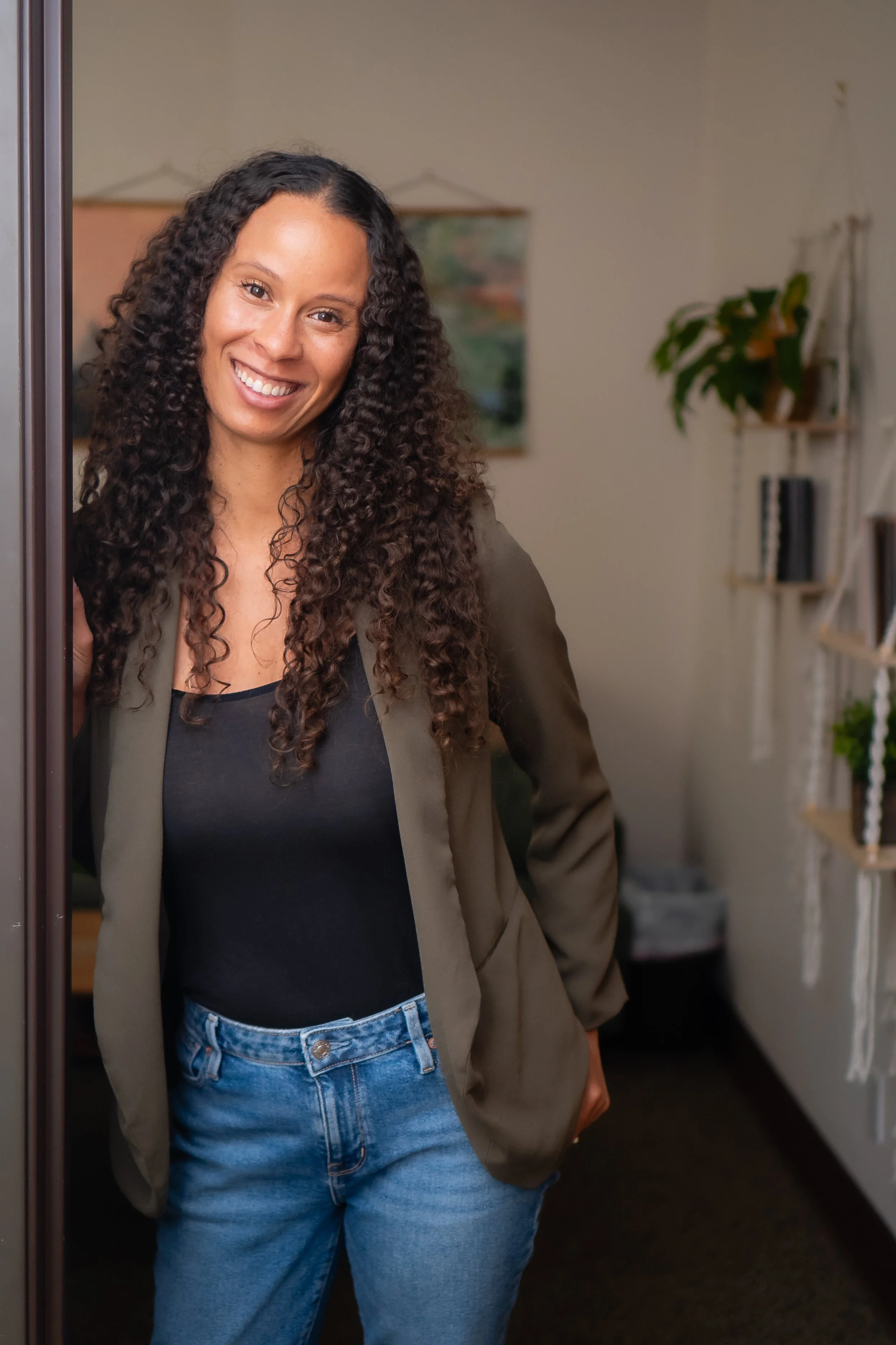Woman with curly hair smiling, wearing a black top and a beige blazer, standing indoors near a mirror, with a background of a wall with a painting and a shelf with plants and books.