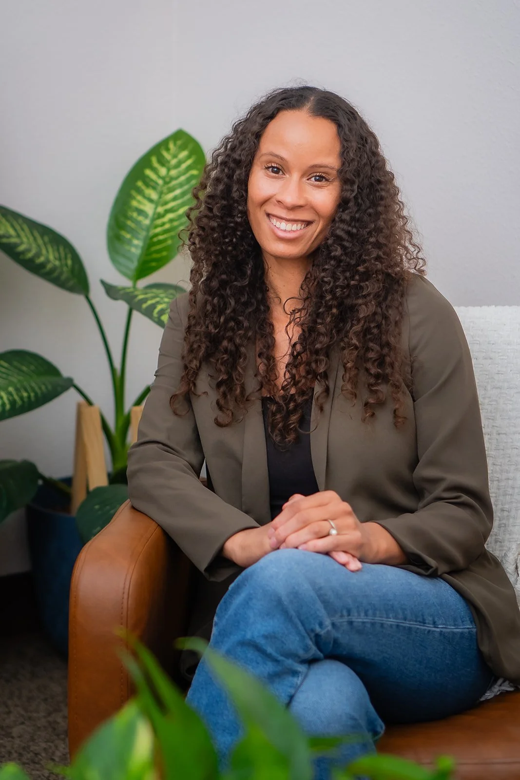 A woman with curly hair smiling, sitting on a tan armchair with a green jacket and blue jeans, indoors with a plant and a light gray wall in the background.
