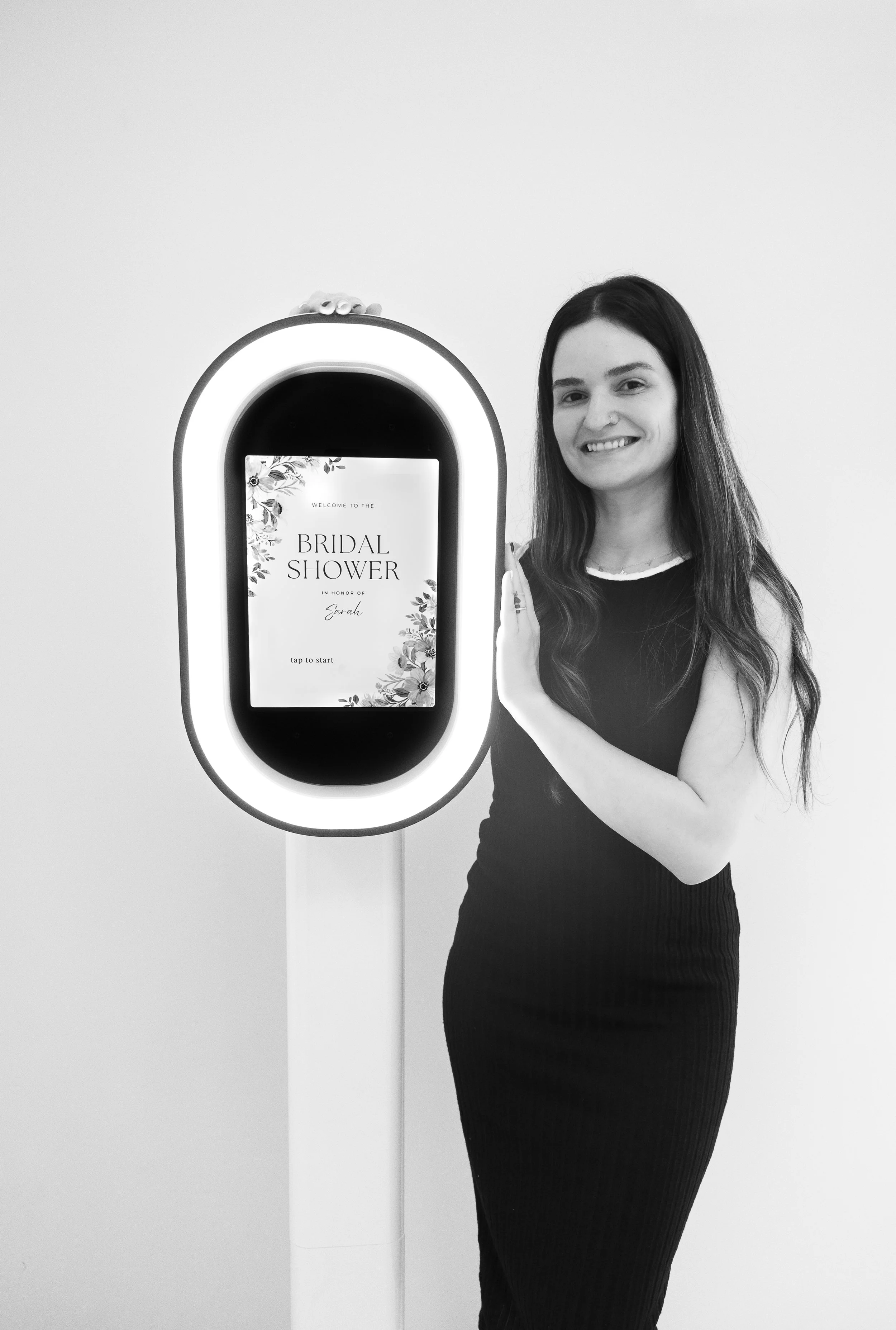A woman with long, wavy hair standing next to a Photo Booth displaying a screen that says 'Welcome to the Bridal Shower in honor of Sarah'. She is smiling and wearing a sleeveless black dress.
