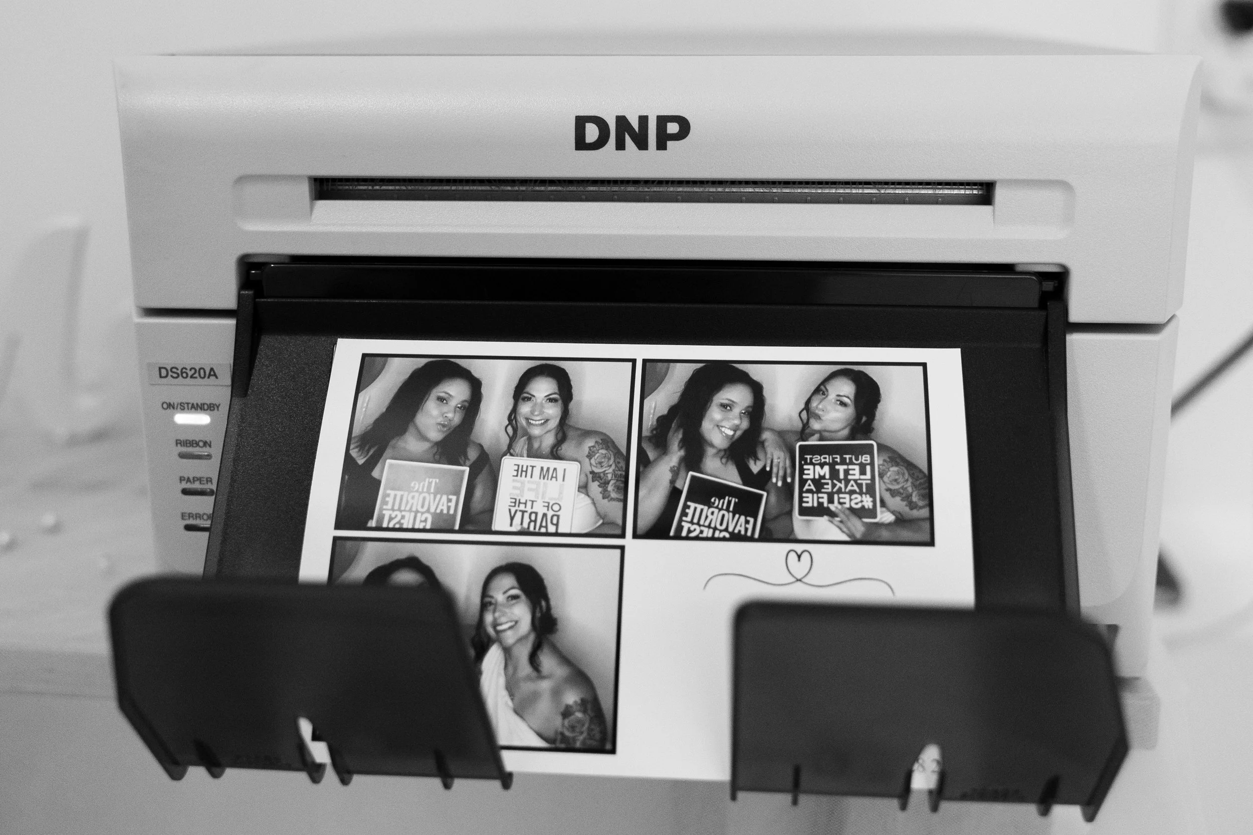 A photo booth printer printing a photo collage featuring four women posing and holding signs, with two women in the first row and one woman in the second row.