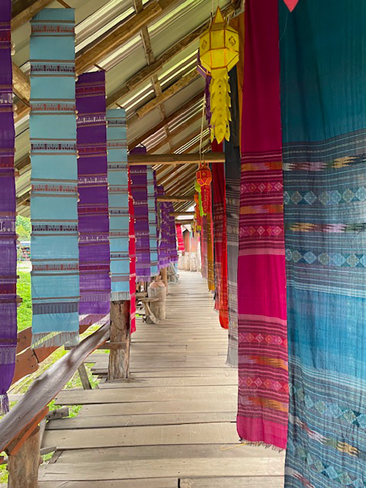 Colorful woven fabrics hanging along a wooden walkway in a traditional market or village setting.