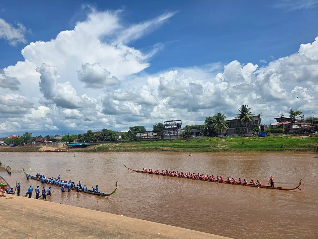 Two traditional long boats racing on a river, each crewed with rowers wearing matching shirts, with one boat in blue and the other in pink. A person stands at the bow of each boat, guiding them. On the riverbank, people are watching the race, with a backdrop of houses, trees, and a sky filled with clouds.